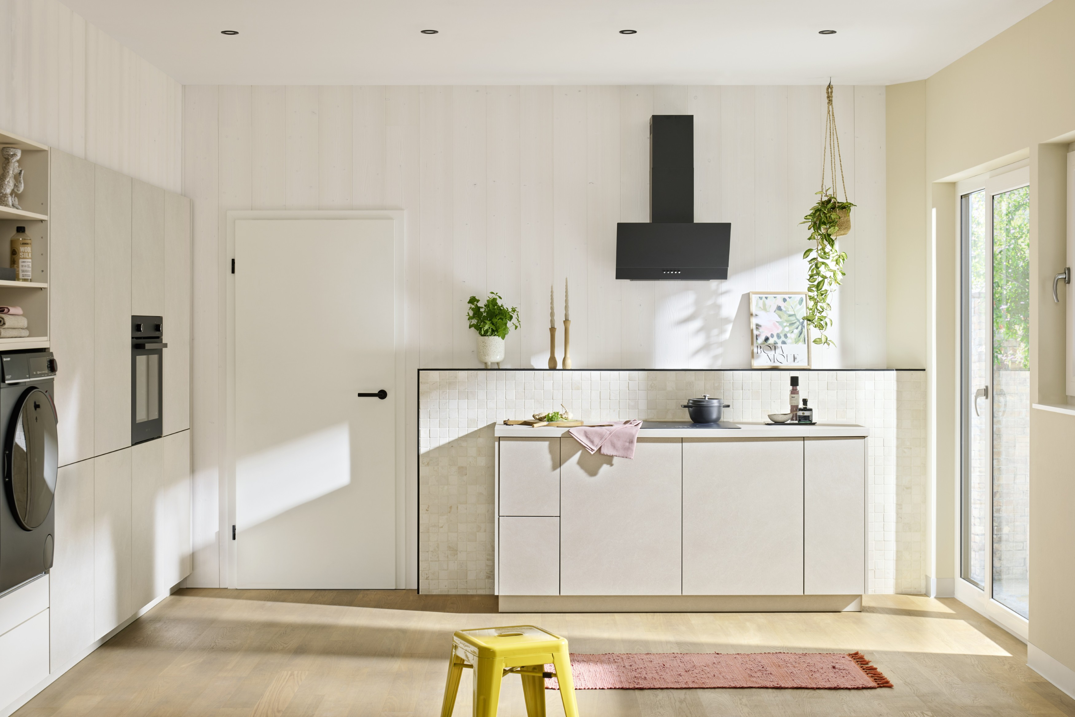 Moderne Küche mit weißen Schränken, schwarzer Dunstabzugshaube, gelbem Hocker, rosa Teppich und Hängepflanze.
