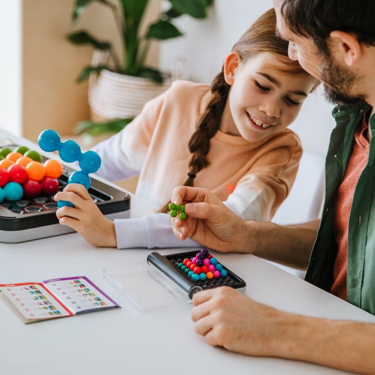 Ein Mädchen und ein Mann spielen ein Puzzlespiel mit bunten Kugeln. Das Mädchen hält ein blaues Teil, der Mann ein grünes.
