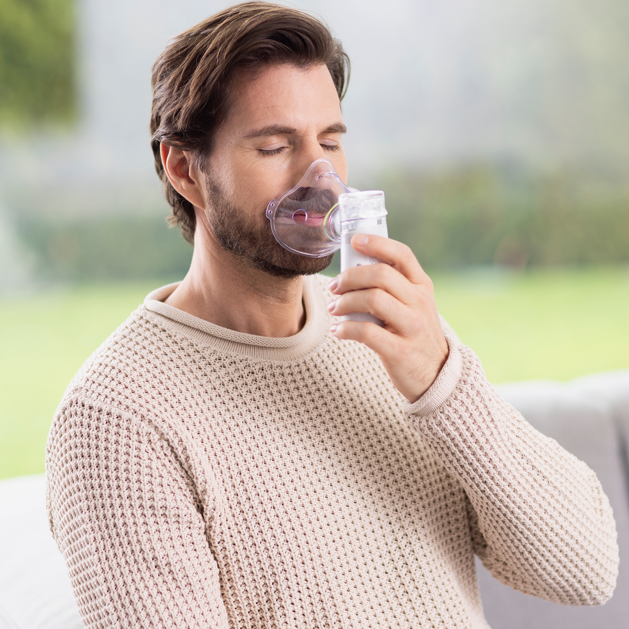 Hombre usando un nebulizador. Lleva una mascarilla y un suéter beige. El fondo es verde borroso.