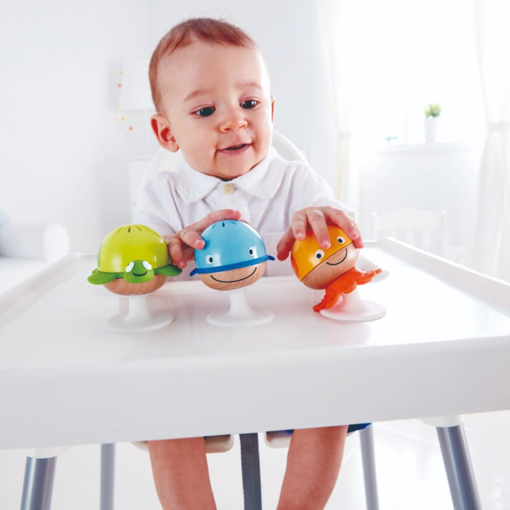 Ein Baby sitzt an einem Tisch und spielt mit drei bunten Spielzeugen. Die Spielzeuge stehen auf weißen Ständern. Das Baby lächelt.