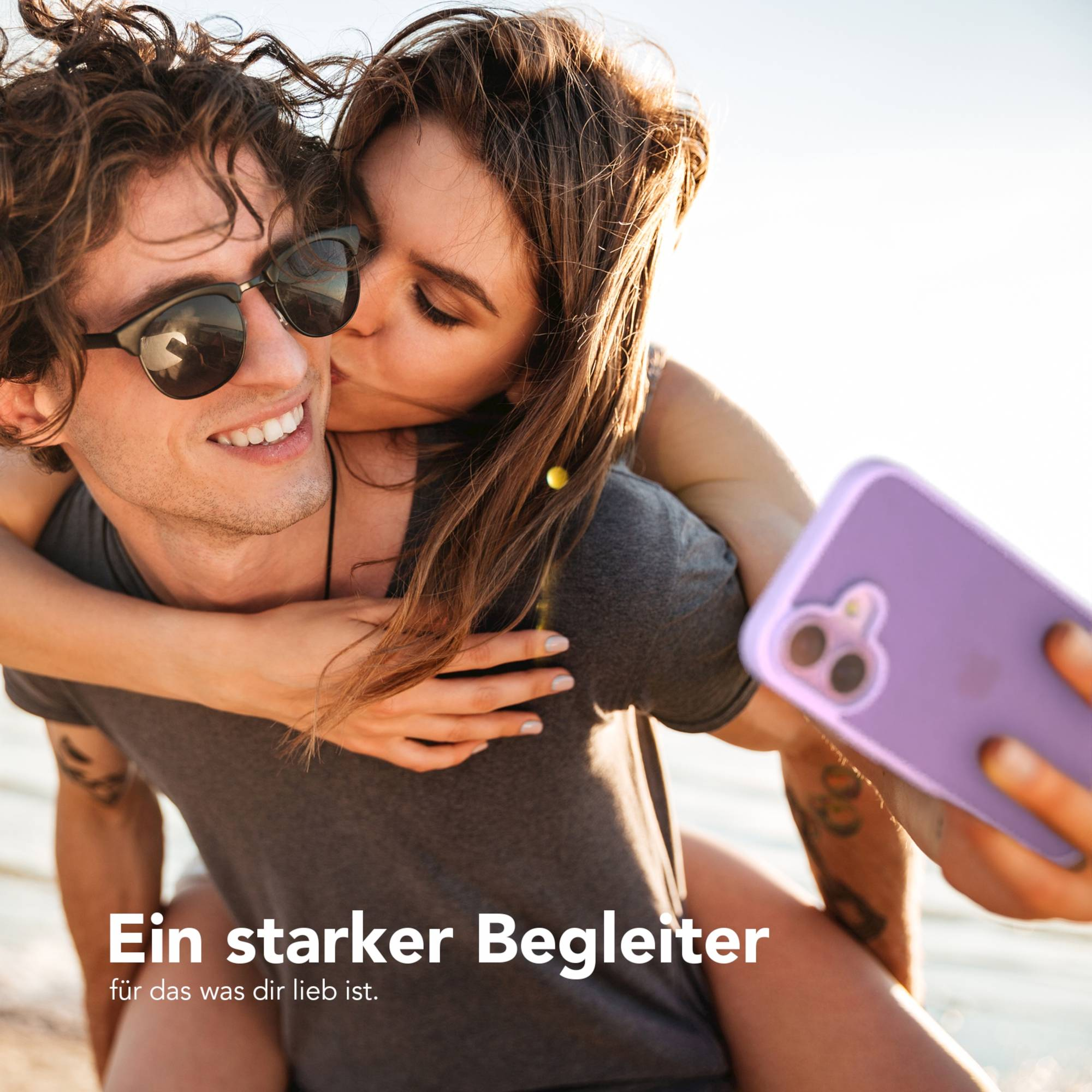 Ein Paar macht ein Selfie. Die Frau küsst die Wange des Mannes. Telefon in lila Hülle. Klarer Himmel im Hintergrund.