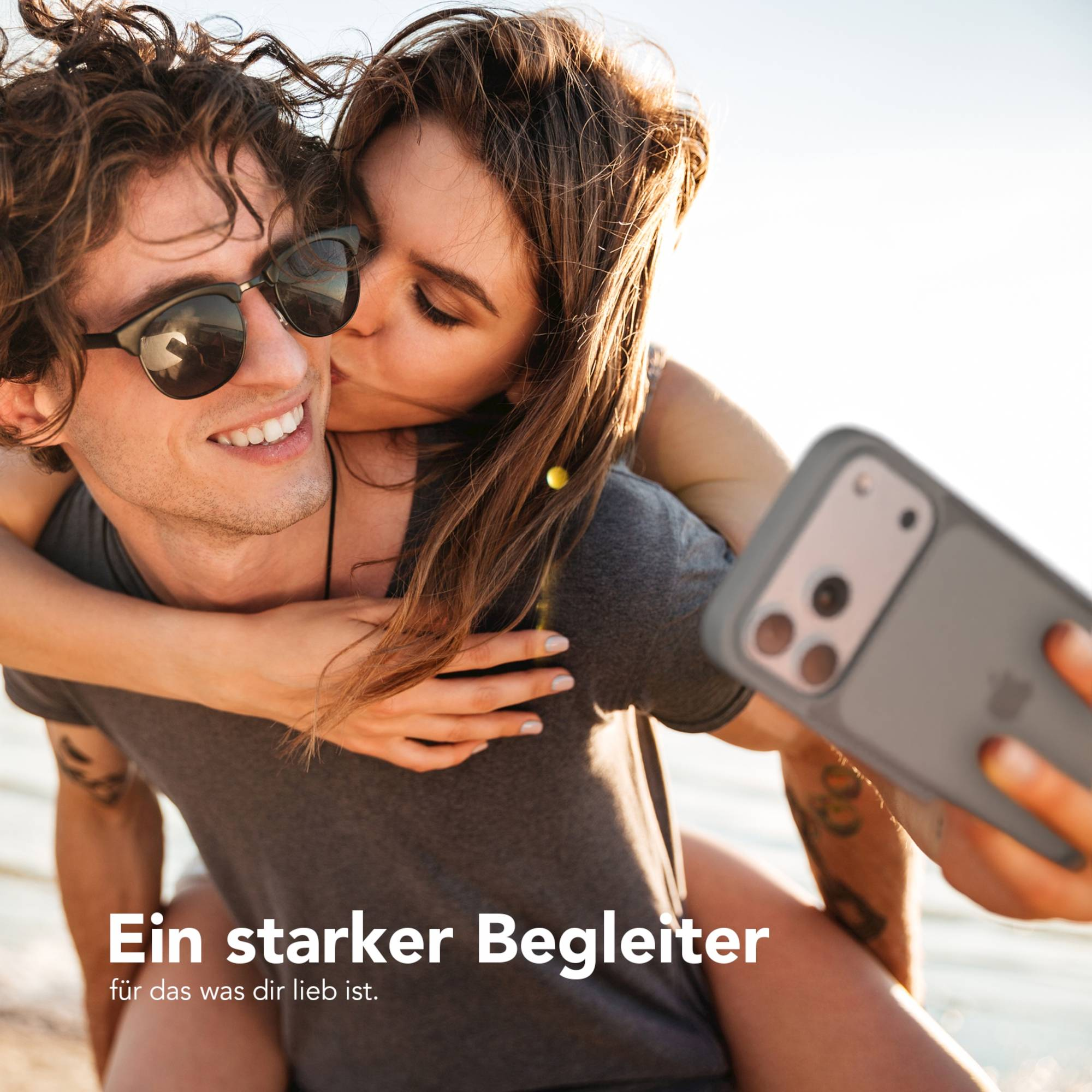 Eine Frau küsst einen Mann auf die Wange. Sie machen ein Selfie. Der Mann trägt eine Sonnenbrille.
