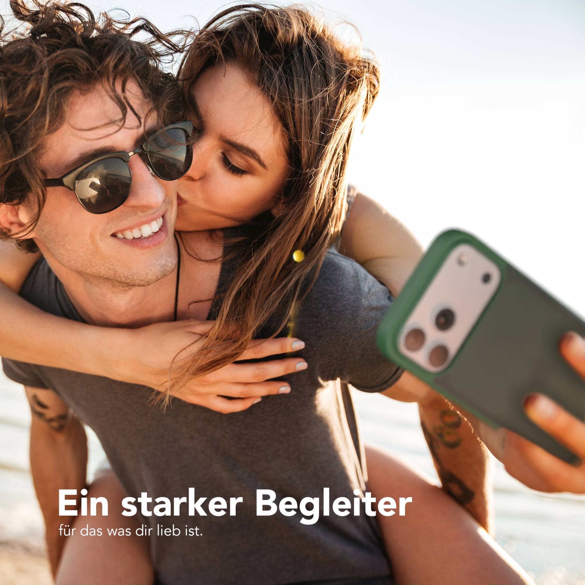 Eine Frau küsst einen Mann mit Sonnenbrille. Sie sind im Freien und halten ein Telefon. Text: Ein starker Begleiter.