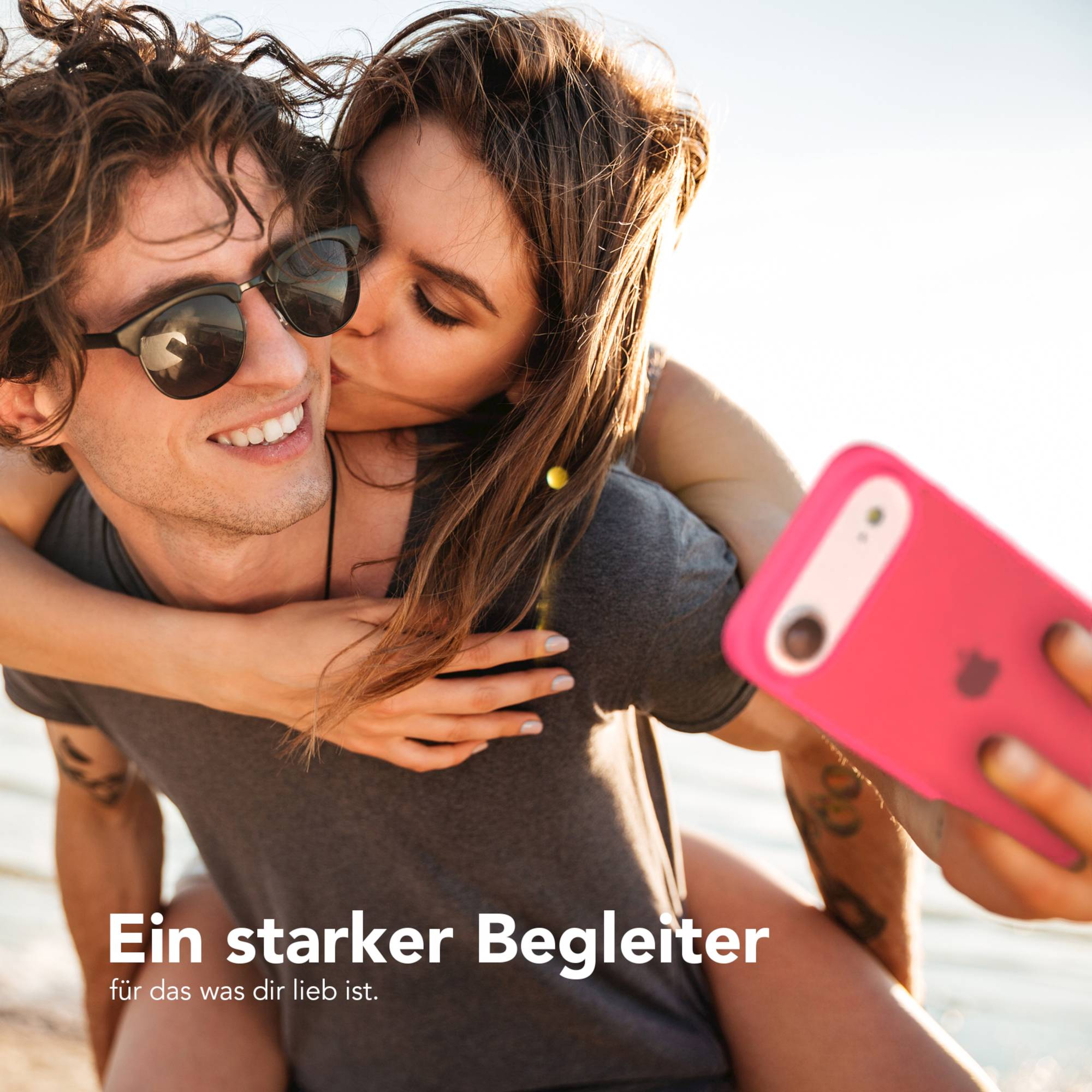 Eine Frau küsst einen Mann mit Sonnenbrille. Sie machen ein Selfie mit einem pinken Telefon.