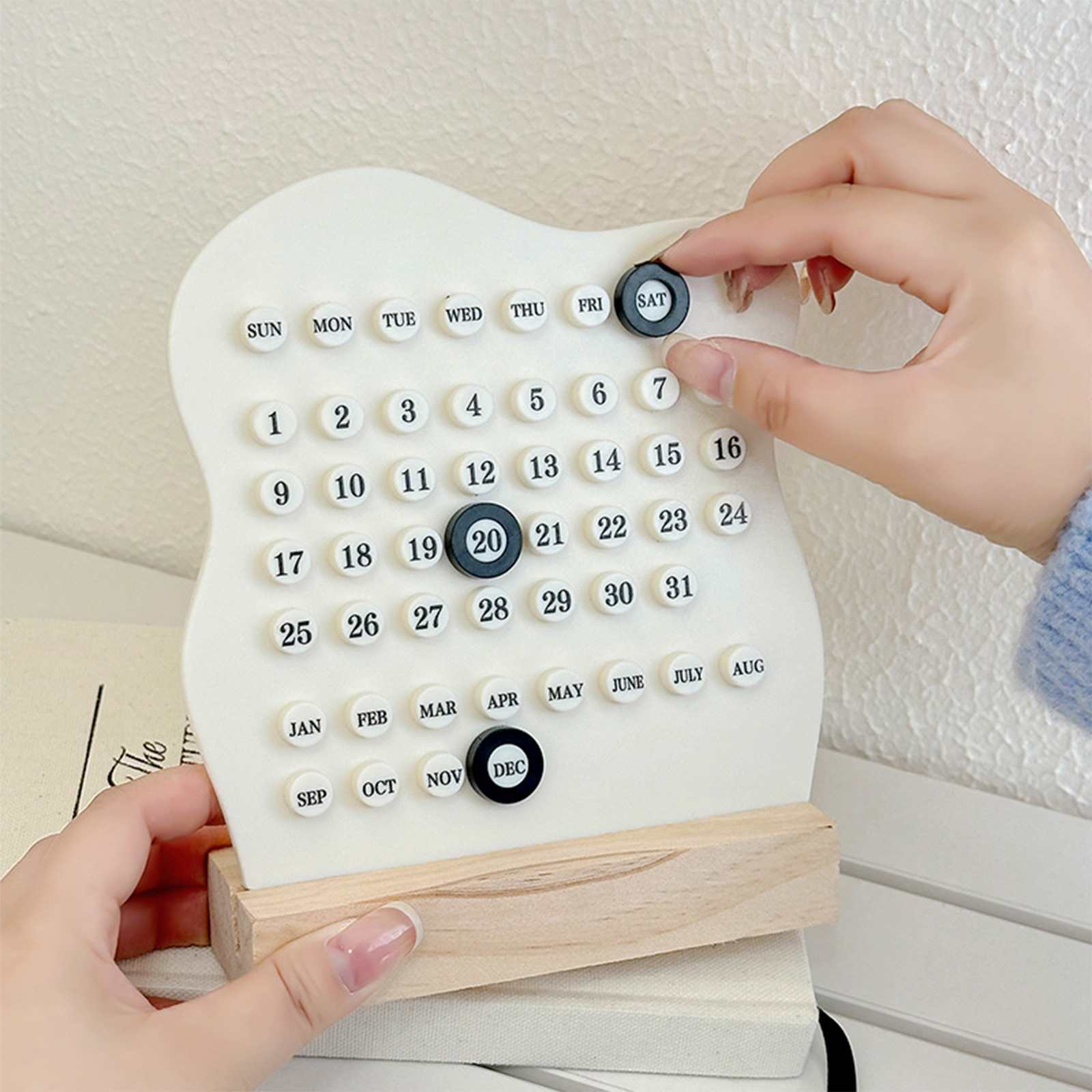 Weißer Kalender mit Holzsockel. Hände passen die Markierung für den Tag 'SAT' an.