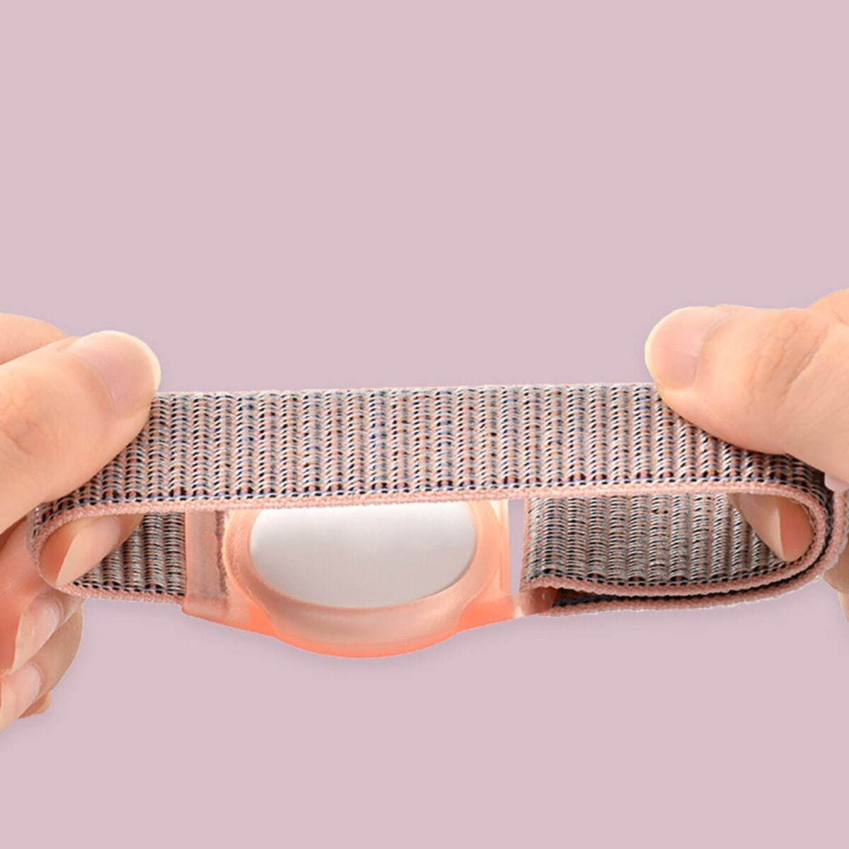 Hände halten ein grau-rosa gewebtes Armband mit einer durchsichtigen rosa Hülle vor rosa Hintergrund.
