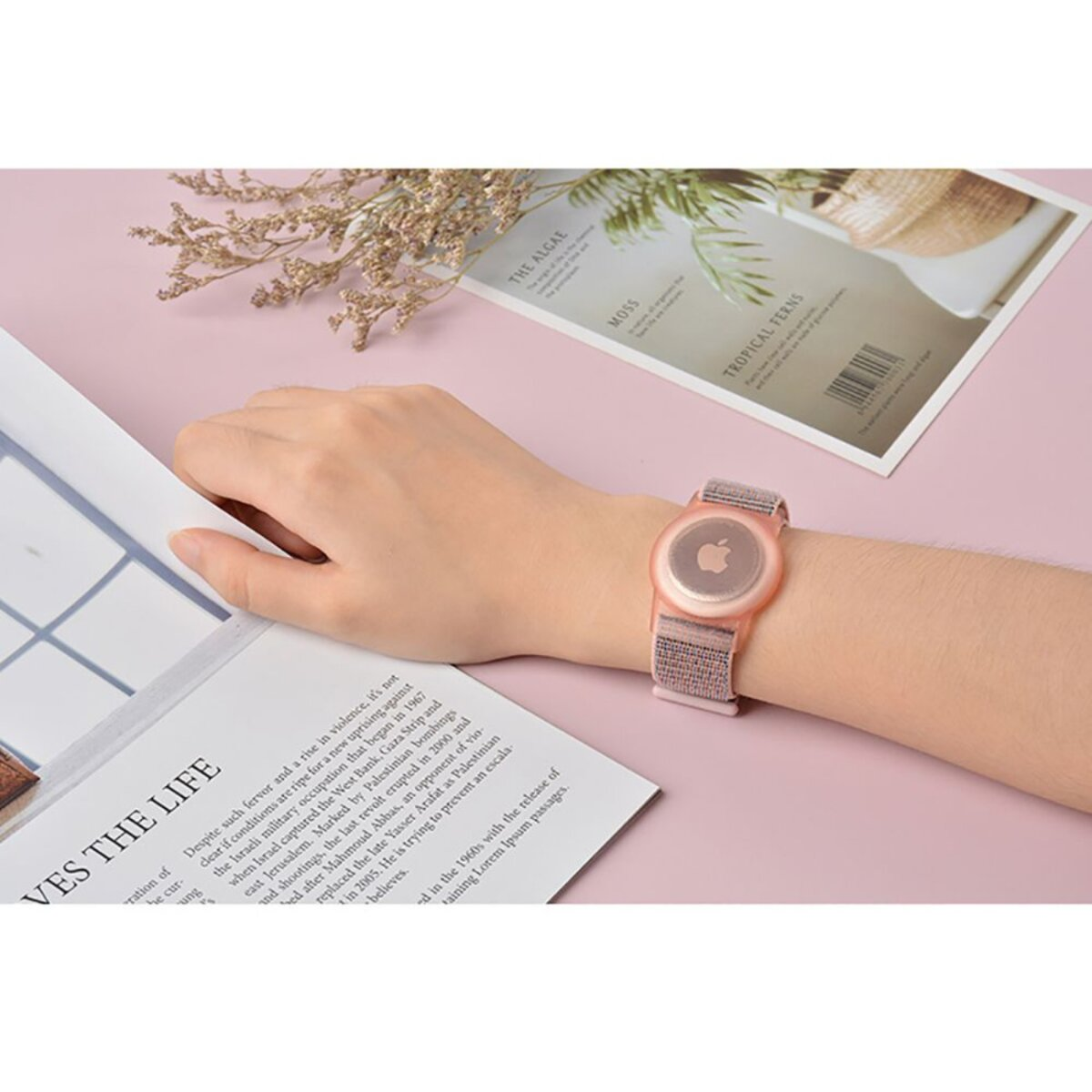 Un poignet avec une montre, à côté d'un magazine avec du texte et une fleur sèche sur fond rose.