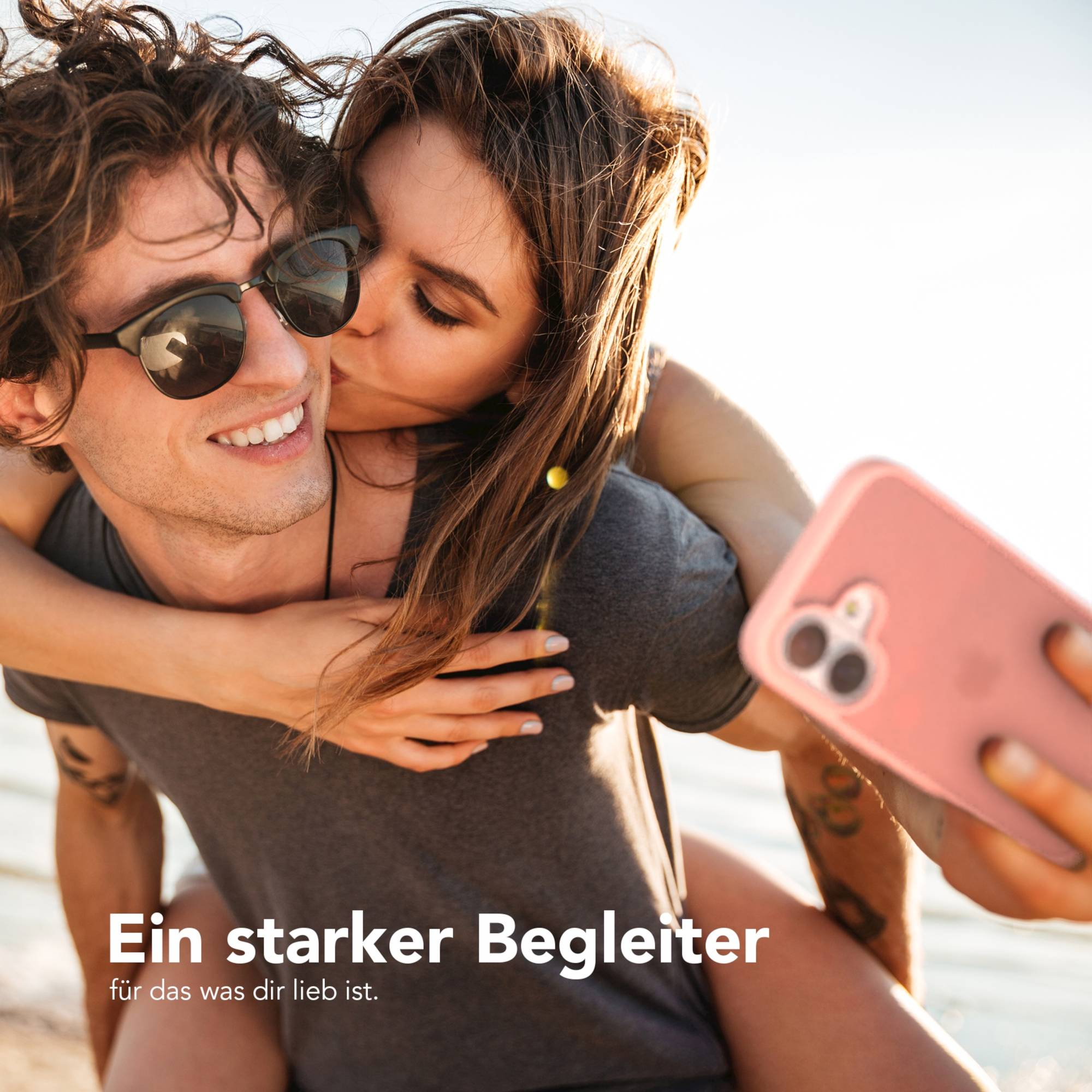 Eine Frau küsst einen Mann auf die Wange. Er trägt eine Sonnenbrille. Sie hält ein Telefon in einem rosafarbenen Gehäuse. Text auf Deutsch.