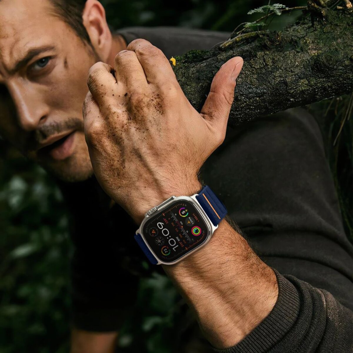 Un homme portant une montre connectée en extérieur, tenant une branche d'arbre. La montre affiche l'heure, les données d'activité. Bracelet bleu foncé. Forêt en arrière-plan.