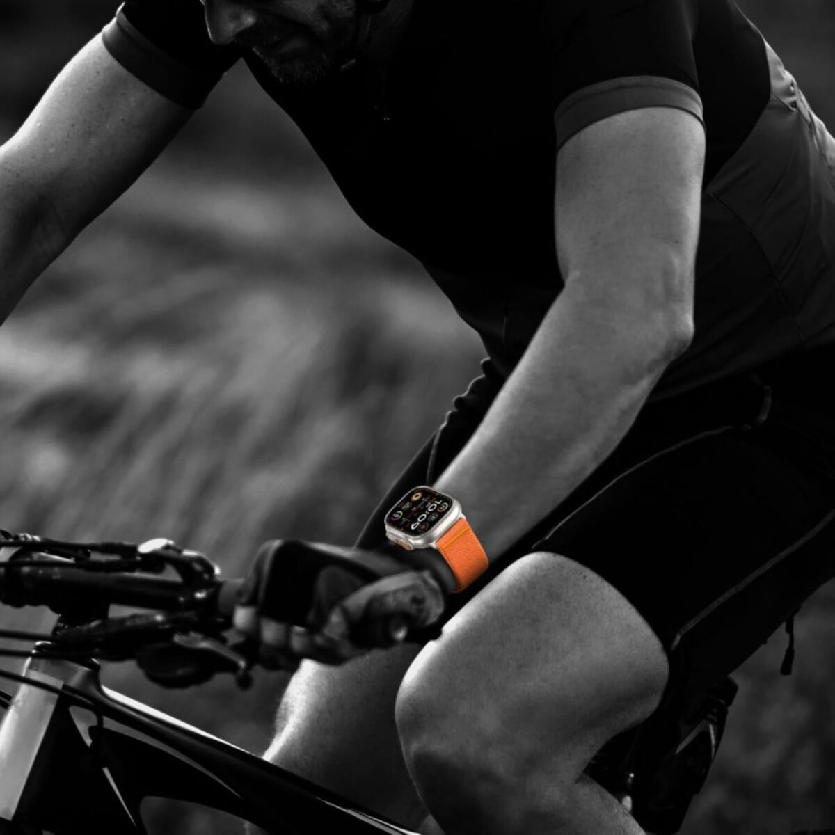 Cycliste avec montre, bracelet orange, faisant du vélo. Fond noir et blanc. Accent sur la montre et le corps du cycliste.