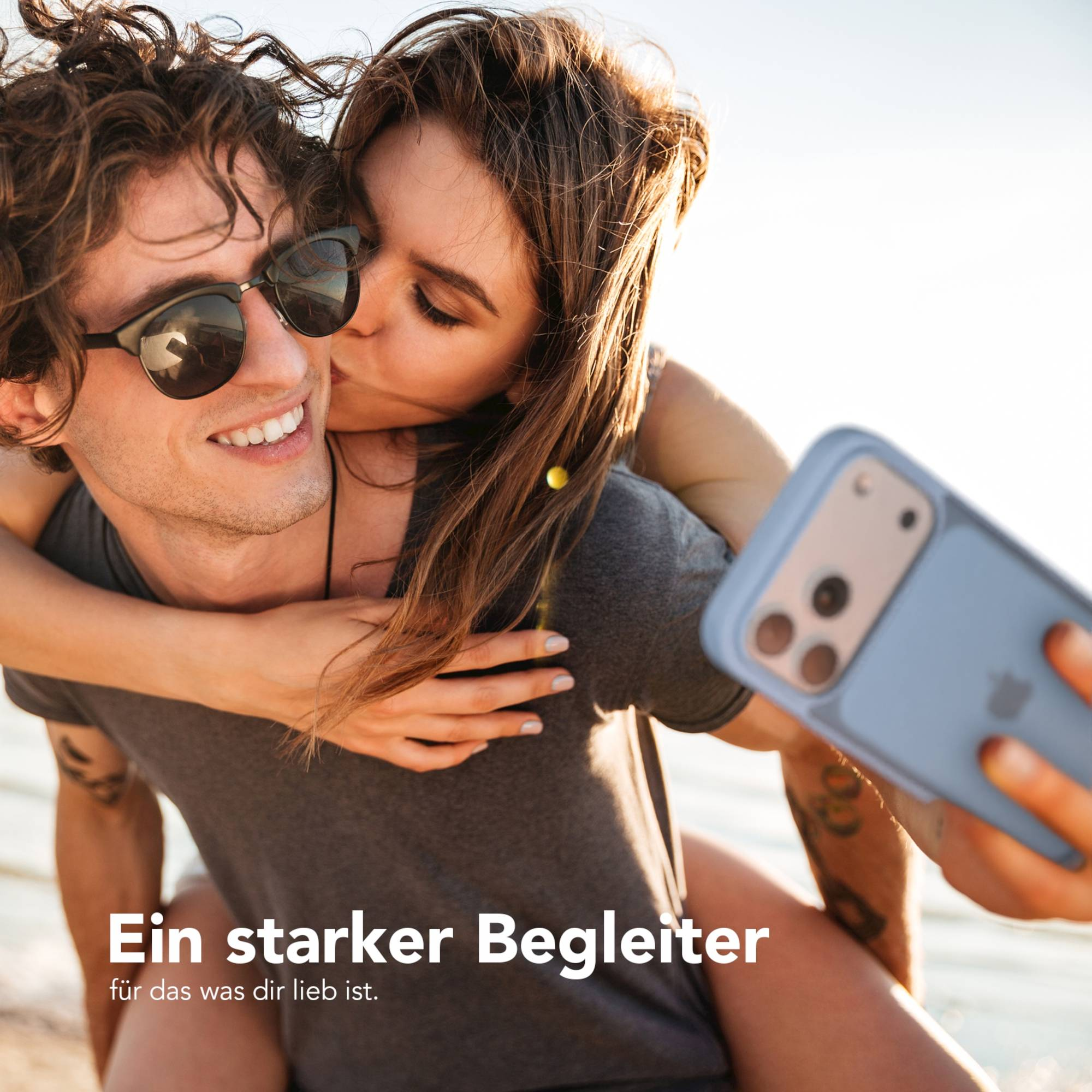 Ein Paar macht ein Selfie. Frau küsst die Wange des Mannes. Mann trägt Sonnenbrille und hält ein Telefon.