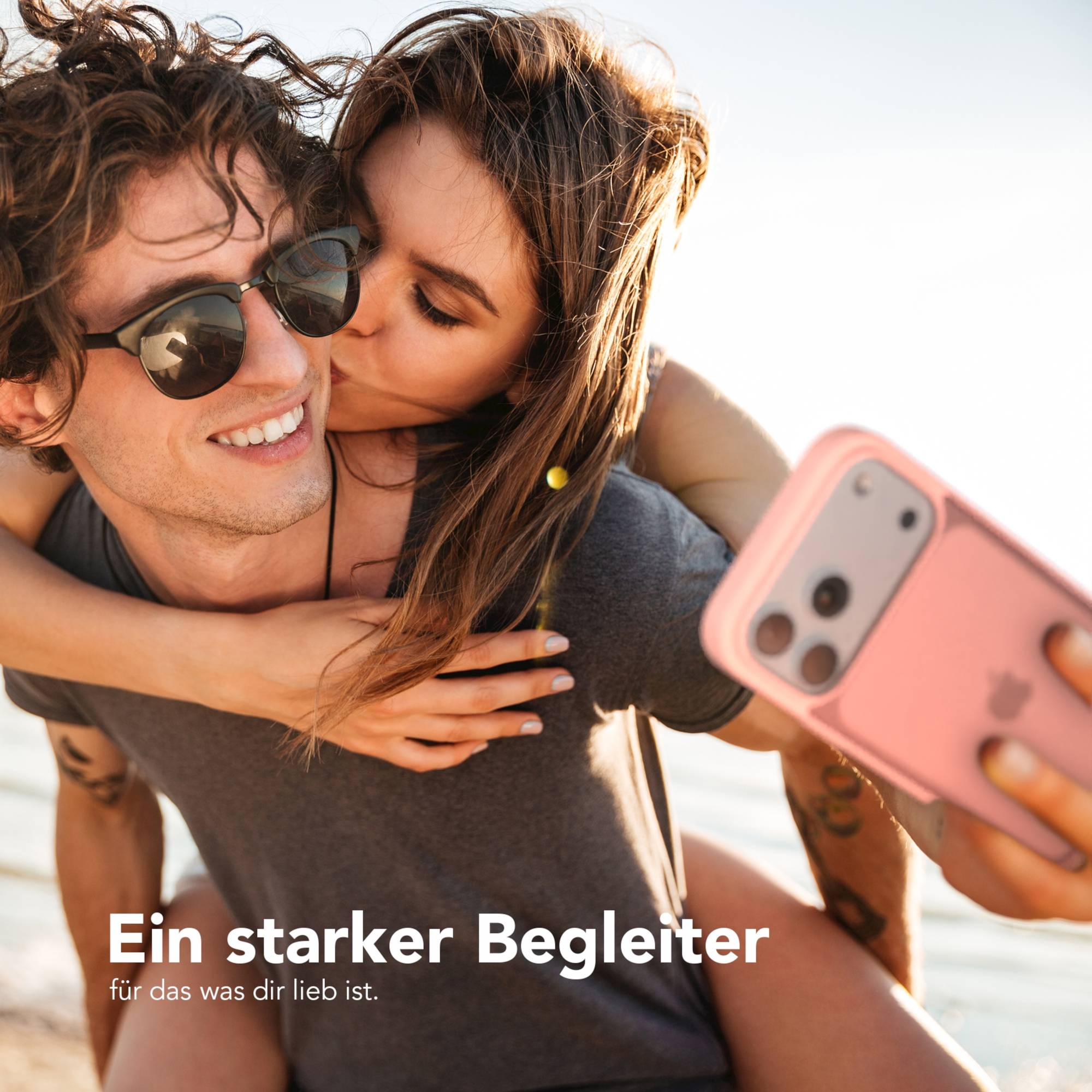 Ein Paar macht ein Selfie mit einer pinken Handyhülle. Die Frau küsst die Wange des Mannes. Beide lächeln im Sonnenlicht.