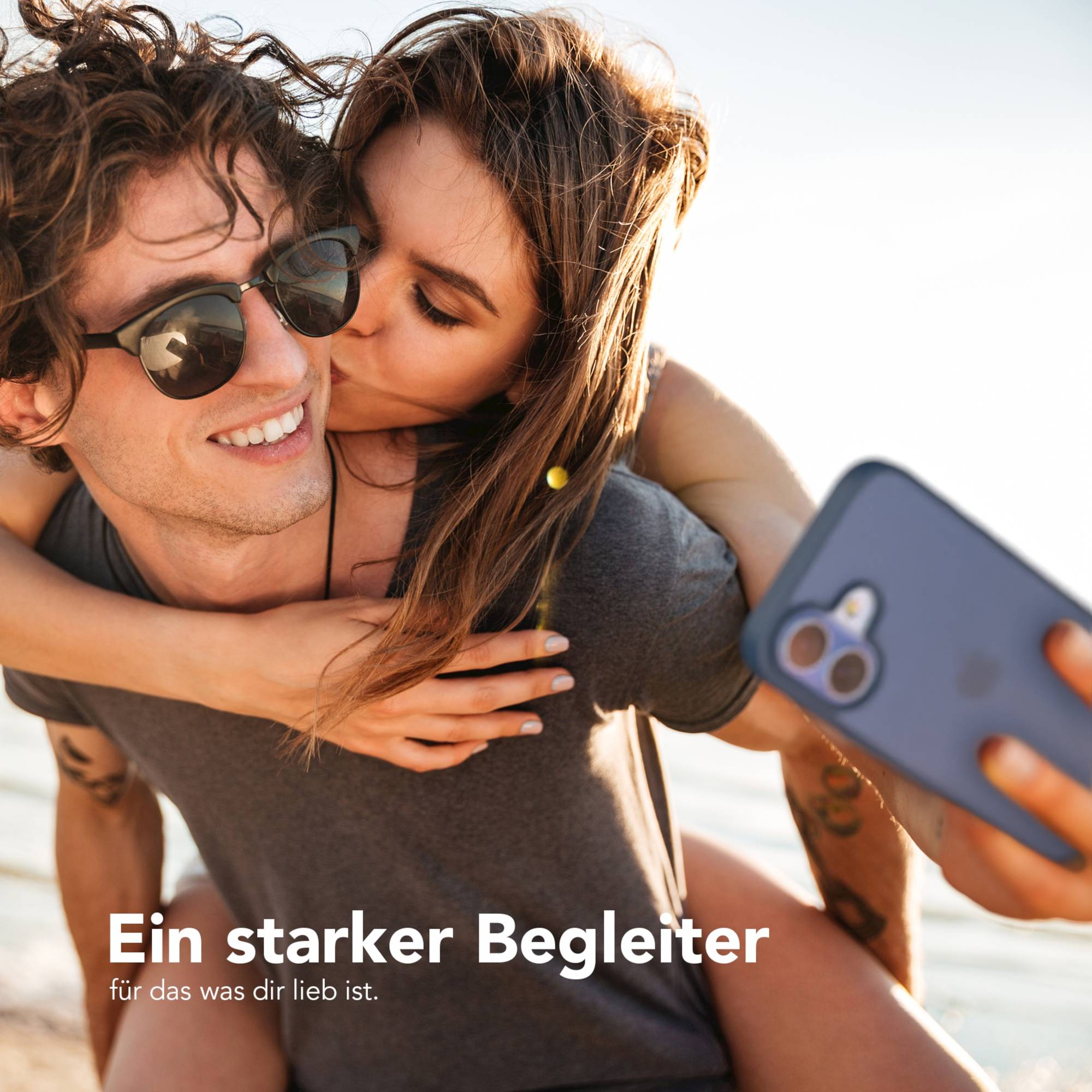 Eine Frau küsst einen Mann auf die Wange. Er trägt eine Sonnenbrille, sie hält ein Telefon. Sie sind draußen.