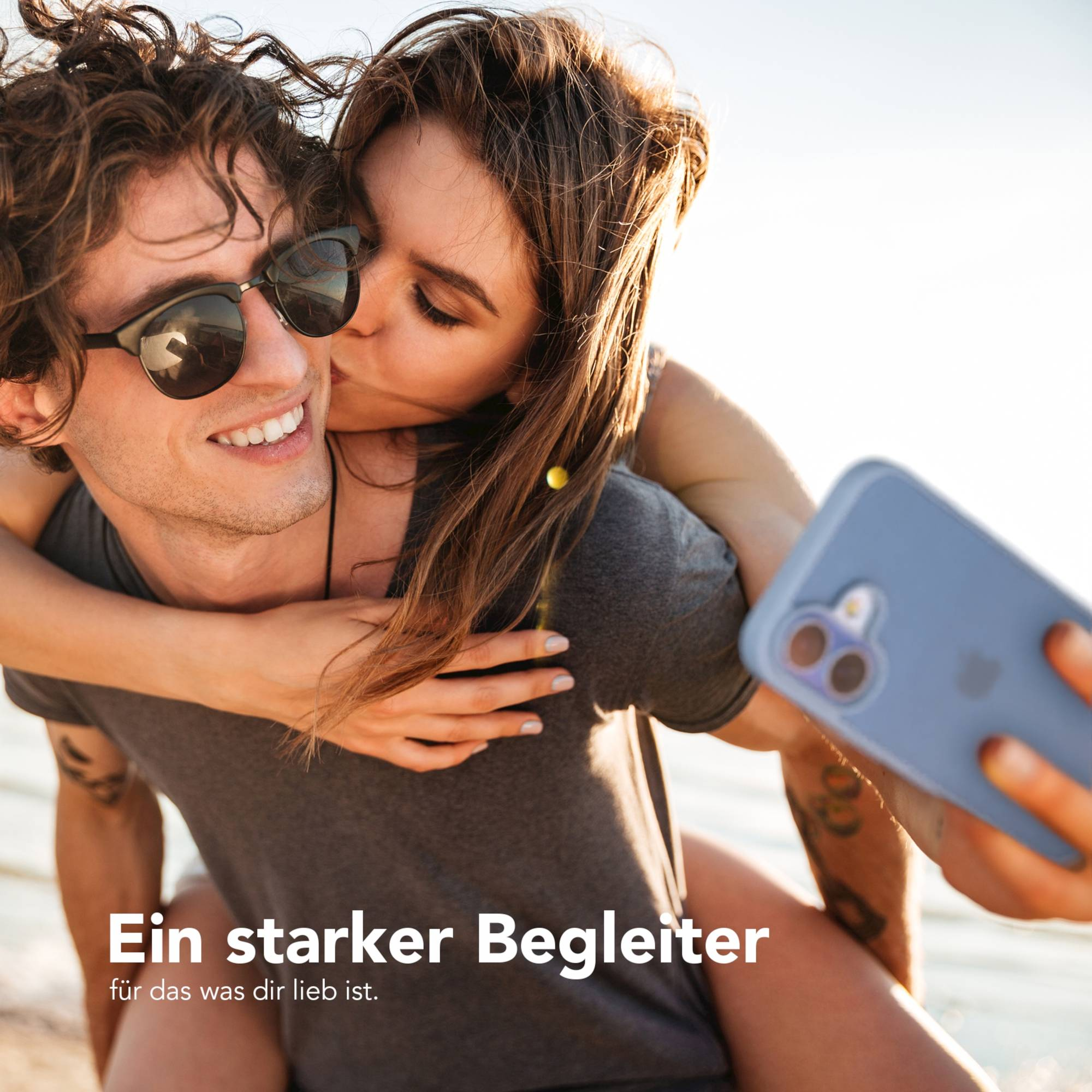 Eine Frau küsst einen Mann auf die Wange, während er ein Telefon hält. Sie sind draußen im Sonnenlicht.