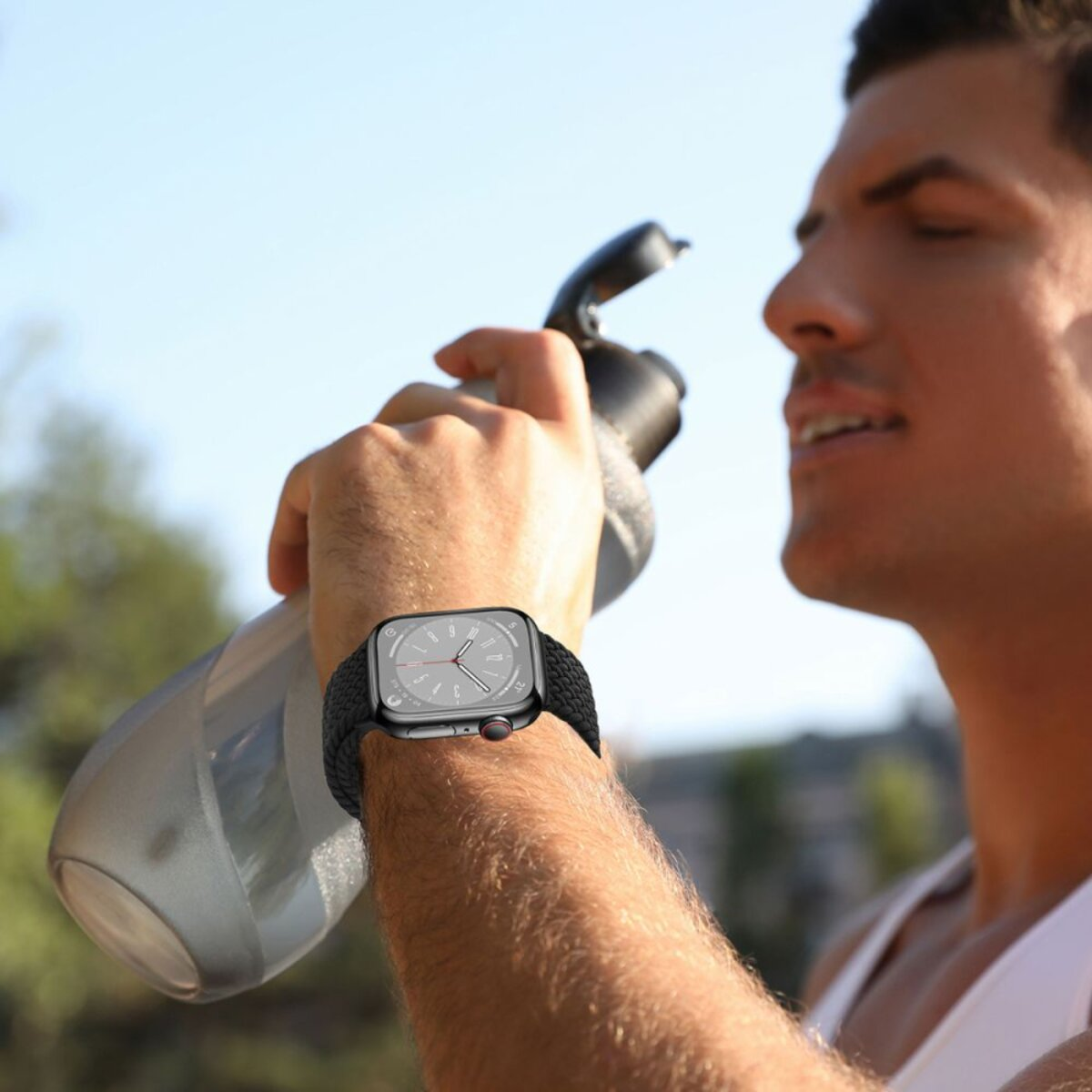 Un homme avec une montre au poignet tient une bouteille d'eau. Il boit à la bouteille, avec un ciel clair en arrière-plan.