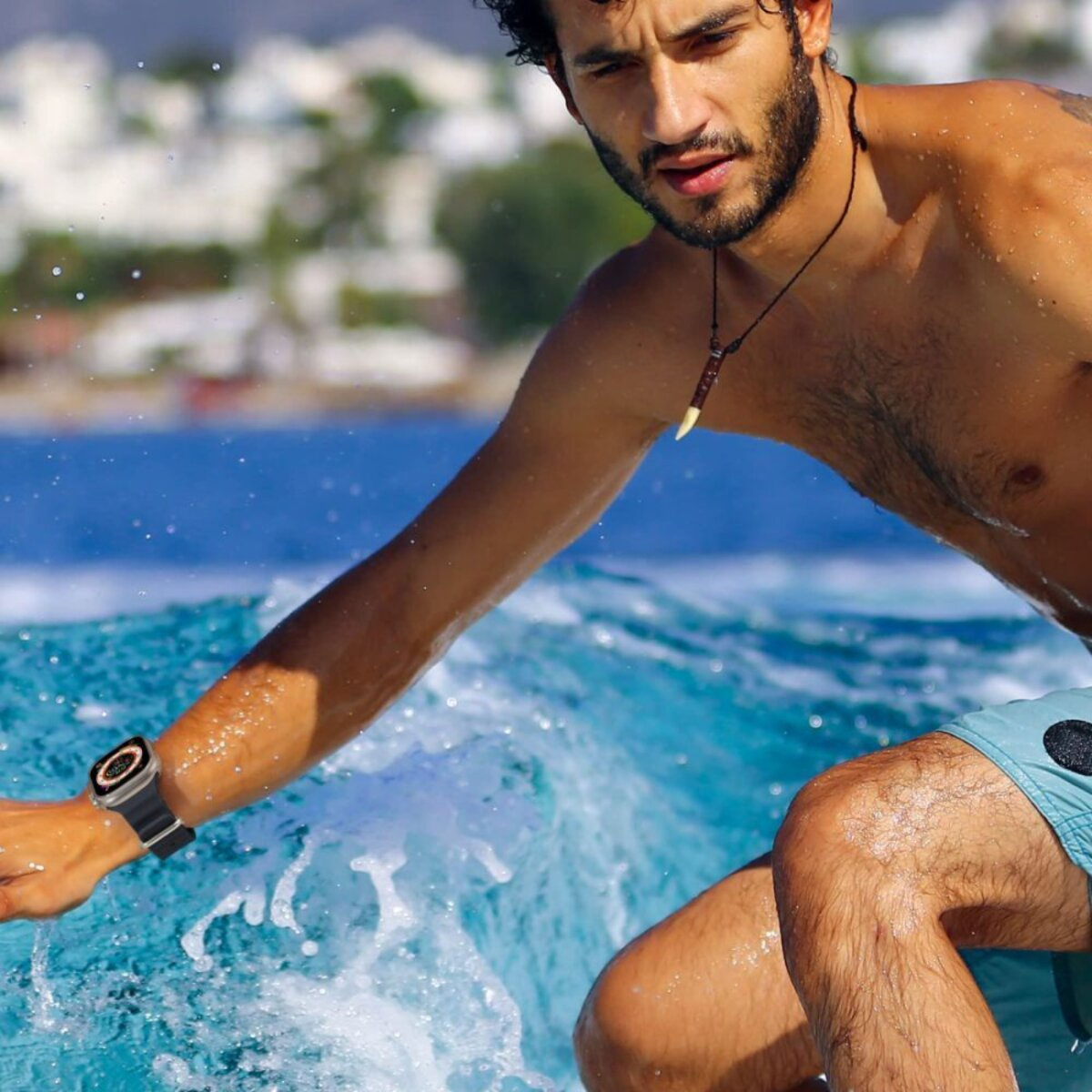 Ein Mann surft. Er trägt eine Uhr und eine Halskette. Das Wasser ist blau und spritzt um ihn herum. Er trägt Badeshorts.