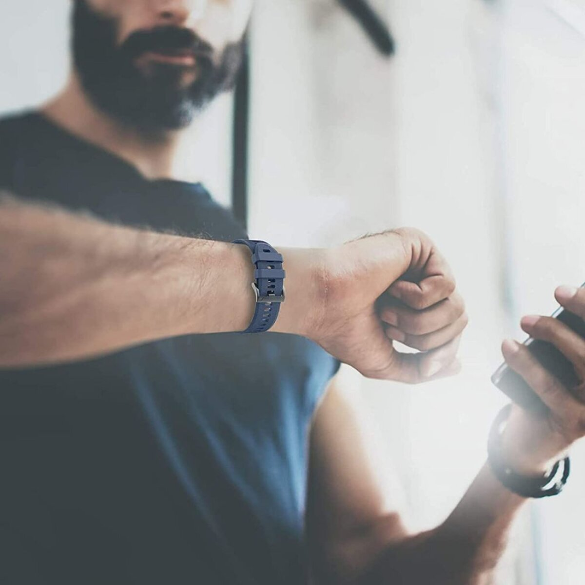 Ein Mann mit blauer Uhr schaut auf sein Handgelenk. Er hält ein Telefon in der anderen Hand. Ein Bart ist sichtbar.