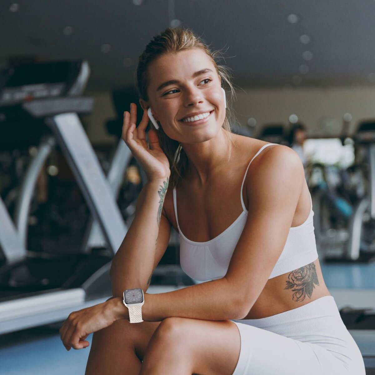 Frau in weißer Sportkleidung im Fitnessstudio, mit Ohrhörern und Smartwatch. Lächelt, mit Tattoo am Arm. Laufbänder im Hintergrund sichtbar.
