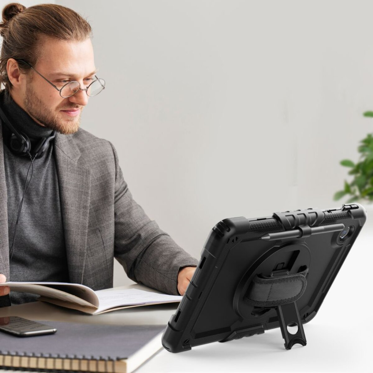 Ein Mann mit Brille liest ein Buch, vor ihm liegt ein schwarzes Tablet. Kopfhörer um den Hals.