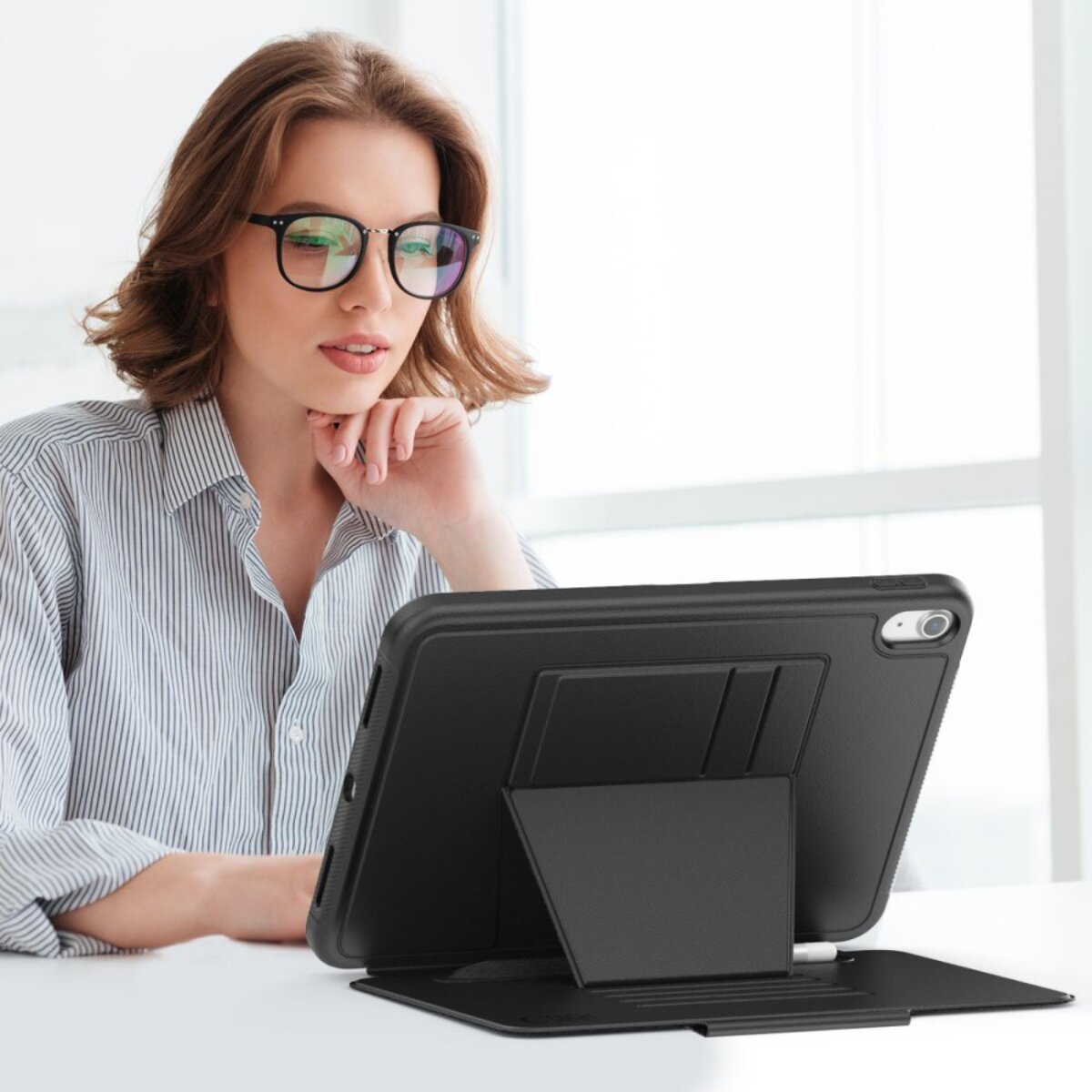 Frau mit Brille schaut auf Tablet. Das Tablet ist in einer schwarzen Hülle mit Ständer. Sie trägt ein gestreiftes Hemd.