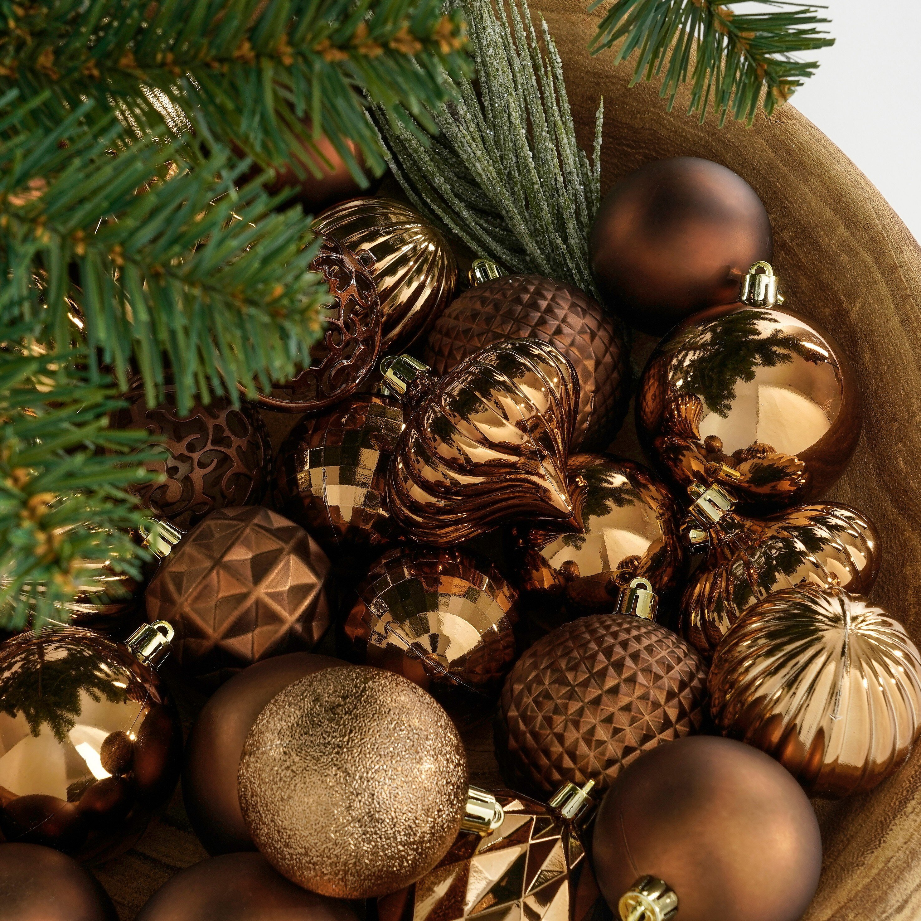 Ornements de Noël bruns dans un bol en bois, aiguilles de pin dans le coin et fond blanc.