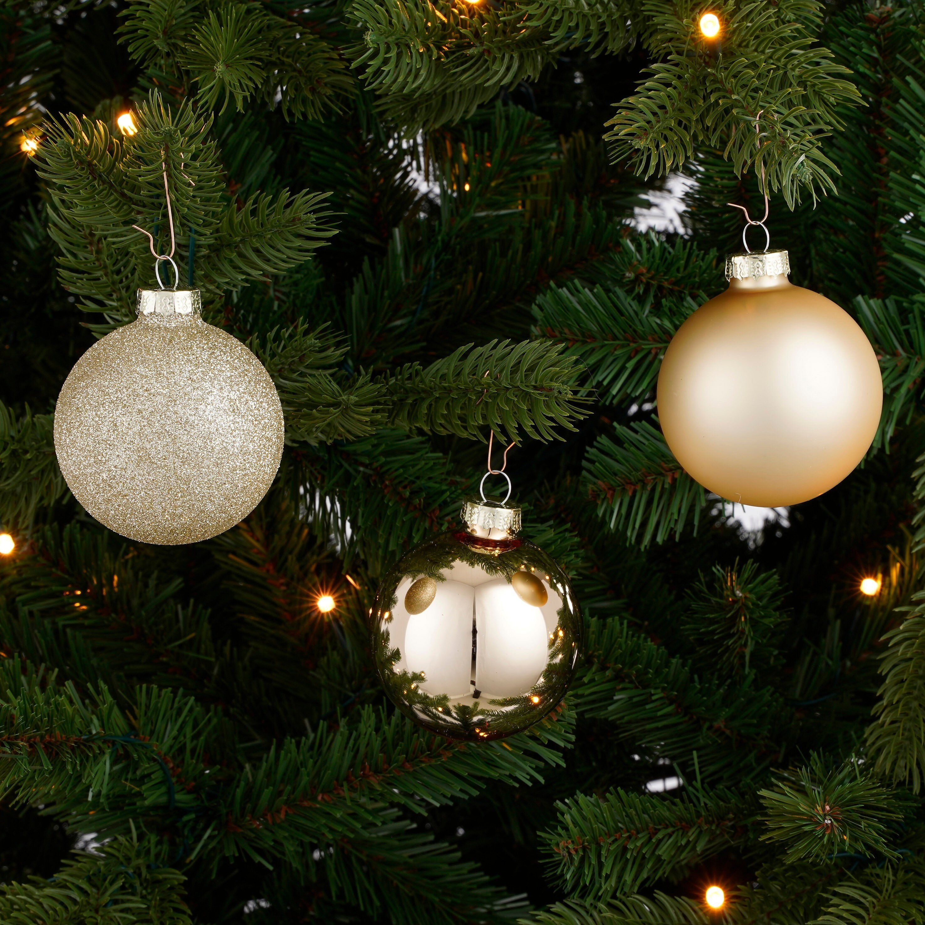 Sapin de Noël avec trois ornements dorés, paillettes, mat et réfléchissants, avec des lumières scintillantes en arrière-plan.
