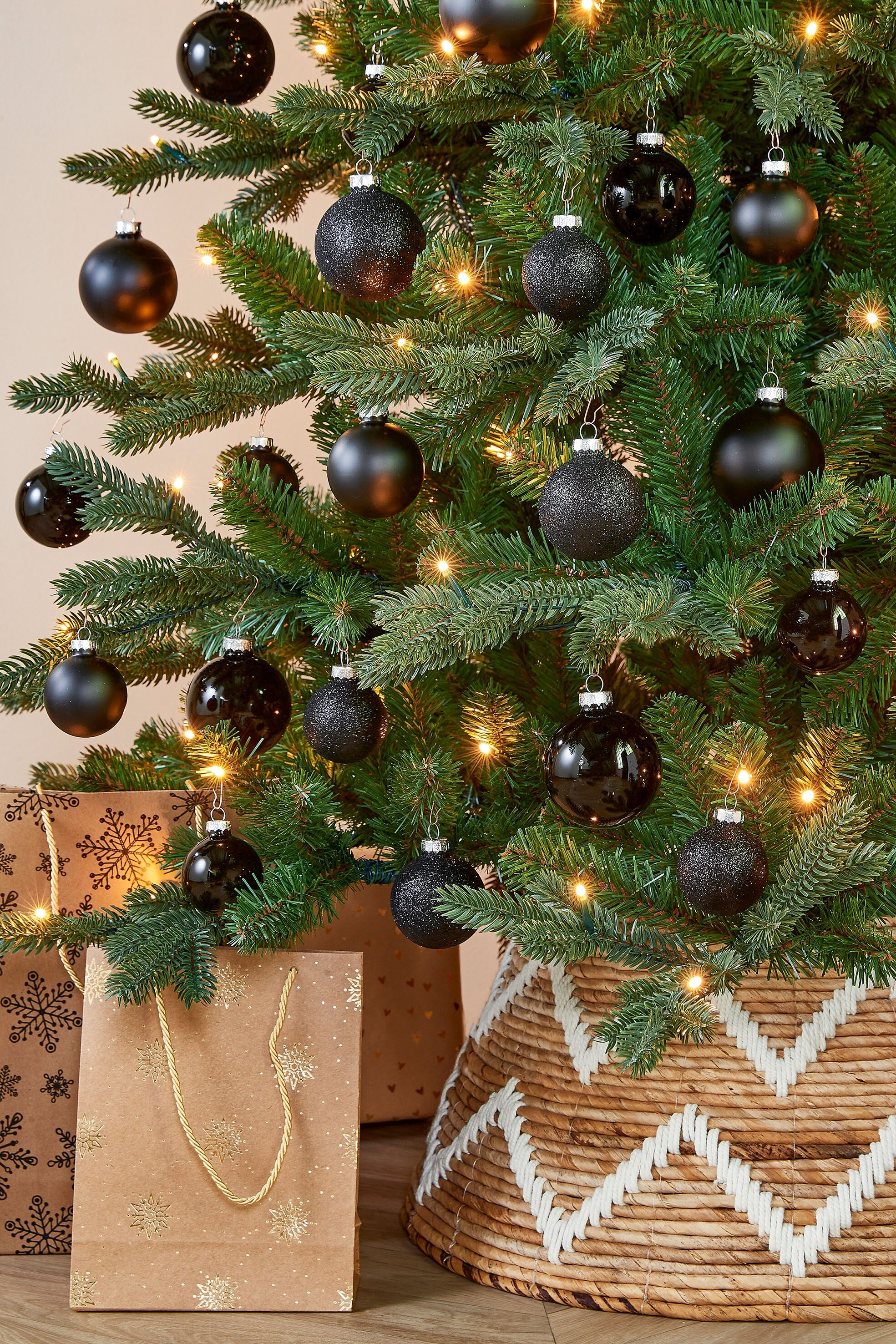 Un sapin de Noël décoré avec des ornements noirs, des lumières et des cadeaux. Un panier tressé entoure la base.