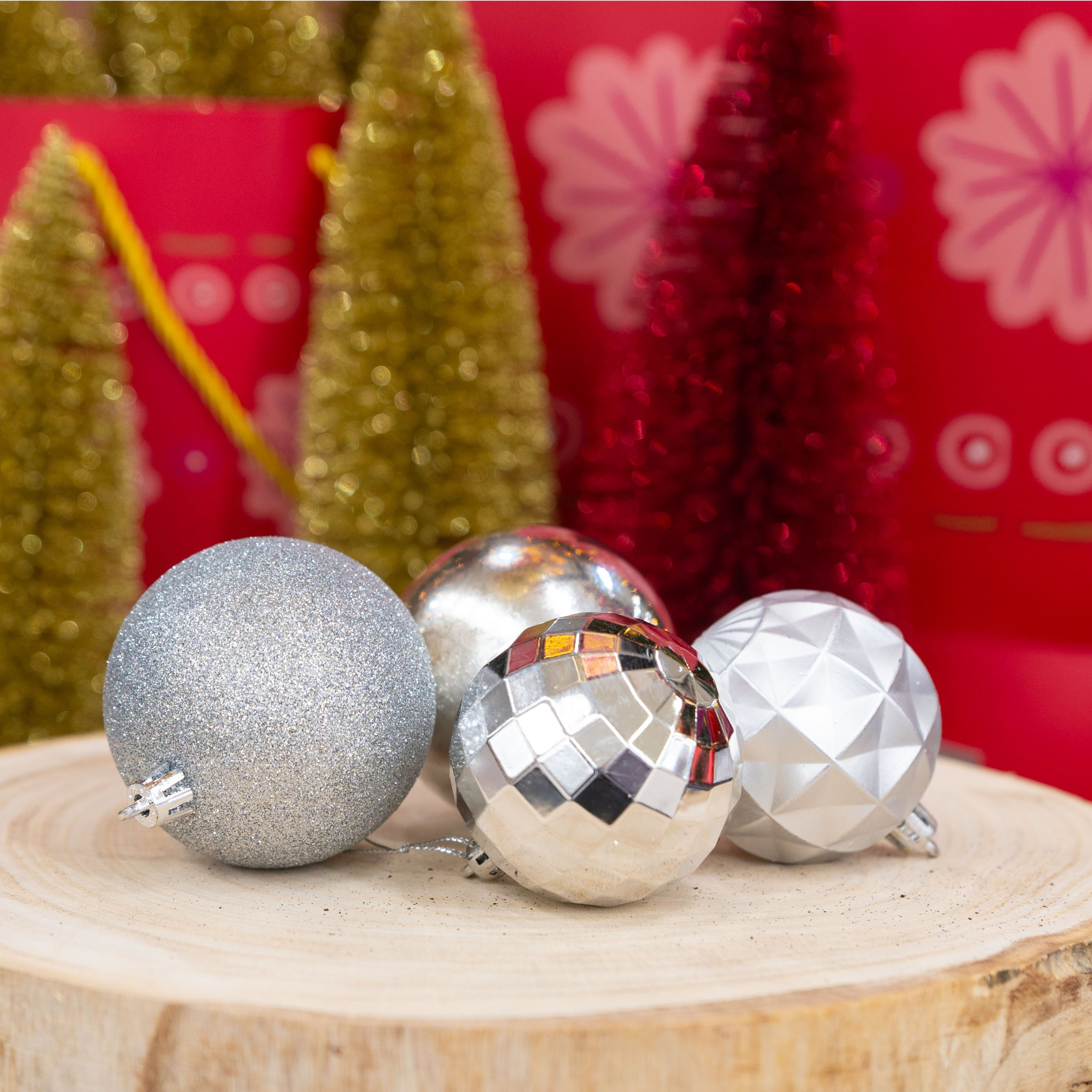 Quatre boules de Noël argentées sur une tranche de bois. Arbres dorés et rouges et sacs cadeaux rouges en arrière-plan.