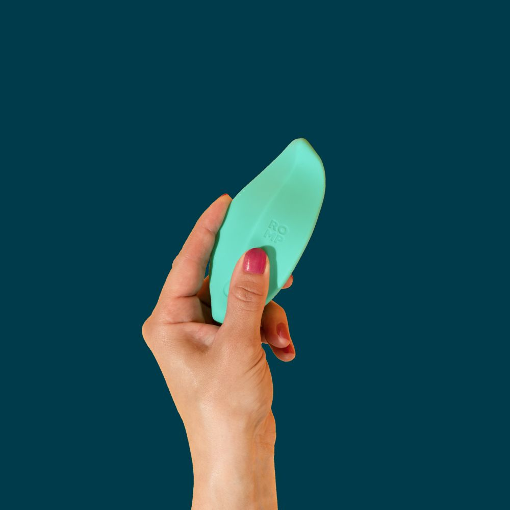 Una mano sostiene un objeto curvo de color verde azulado con las letras 'RO MP'. El fondo es de color verde azulado oscuro.