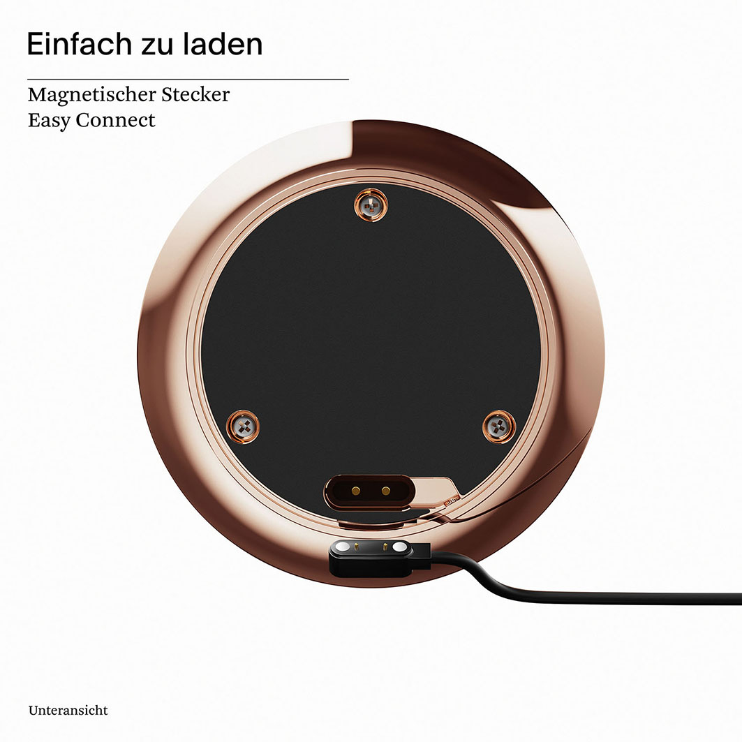 Die Unterseite eines roségoldenen, kreisförmigen Geräts mit schwarzer Mitte. Drei Schrauben sind sichtbar. Ein schwarzes Ladegerät ist eingesteckt.