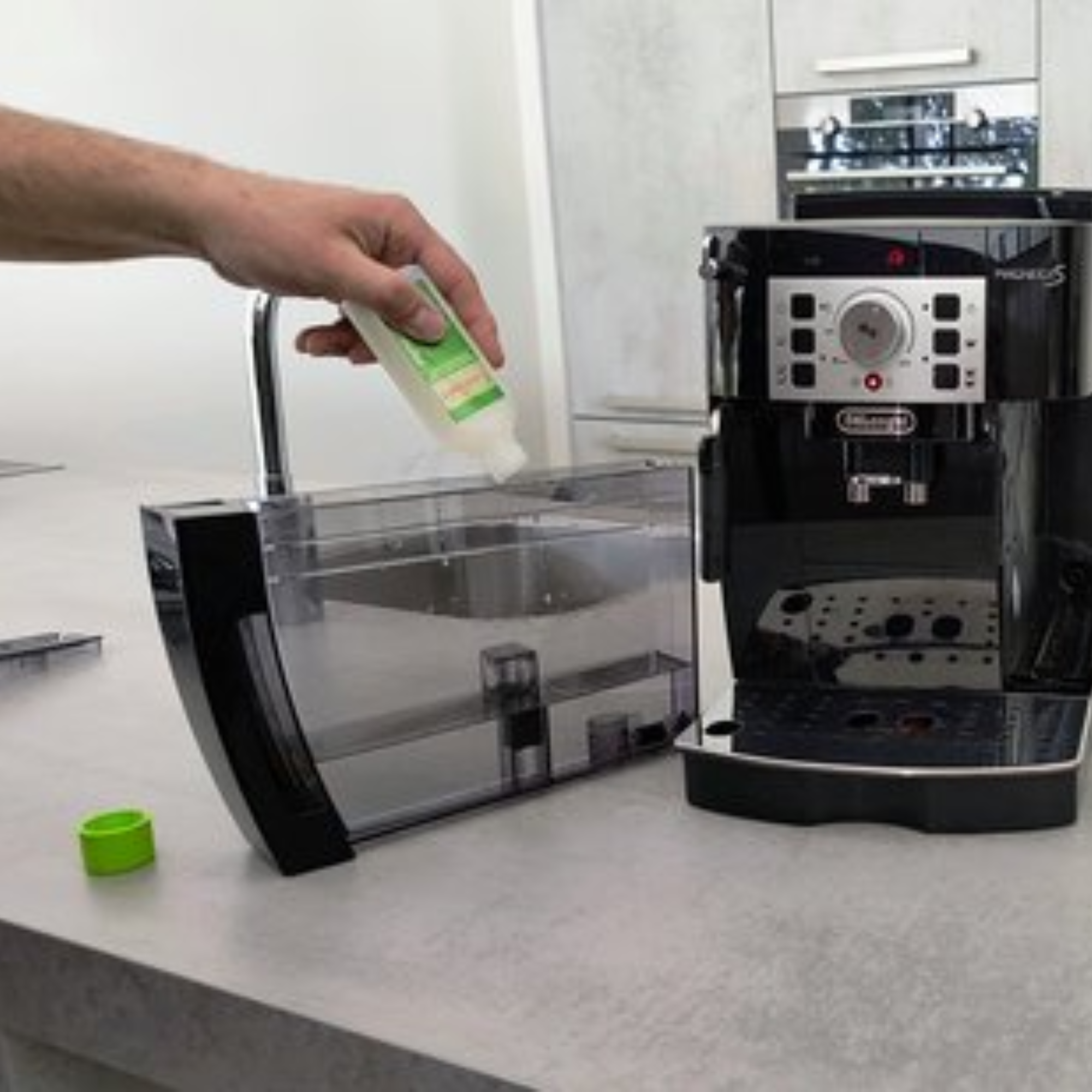 A person pours liquid from a bottle into a coffee machine's water tank. A green cap sits nearby.