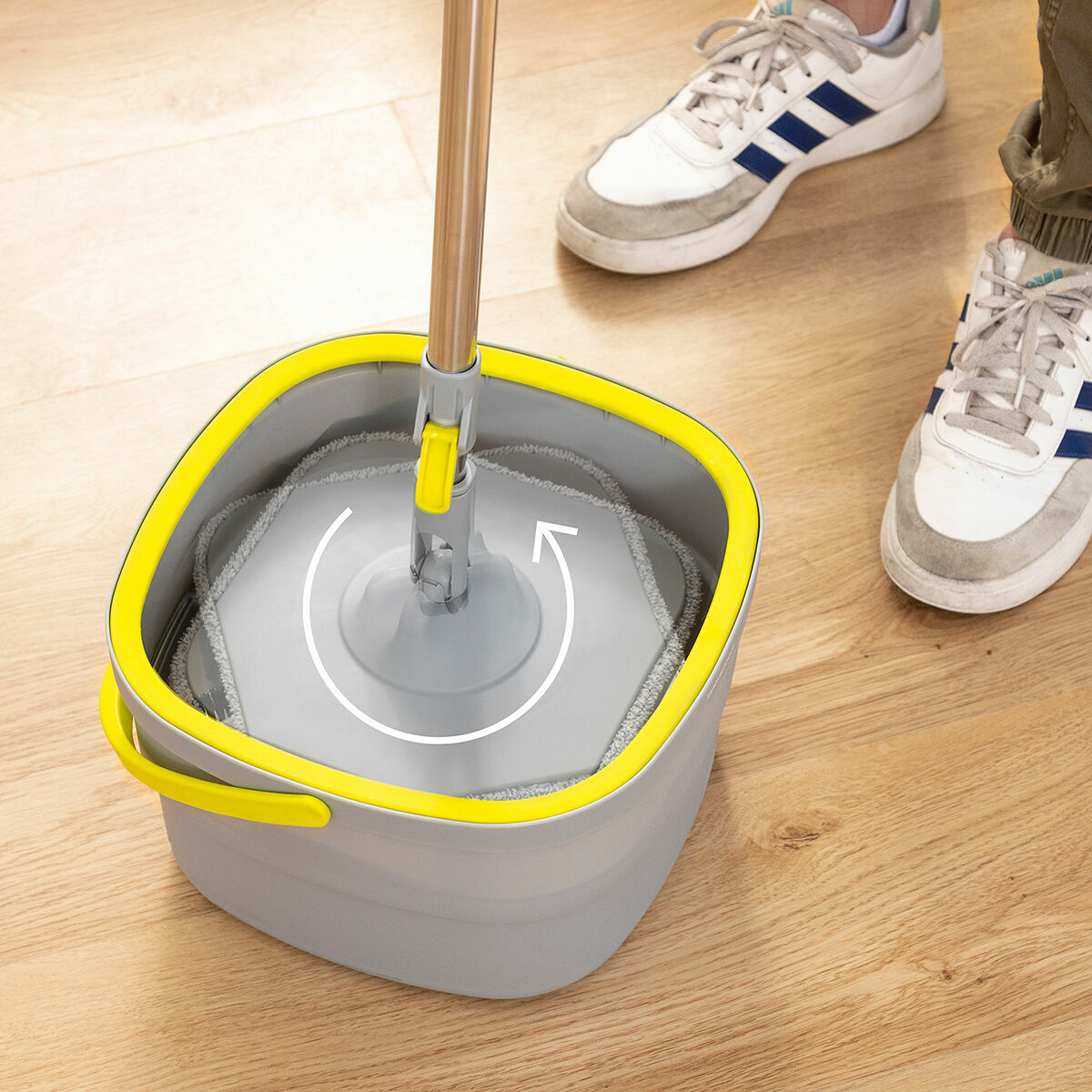 Un secchio per mocio grigio e giallo con un mocio all'interno, una persona sta usando il mocio. Scarpe da ginnastica bianche sullo sfondo.