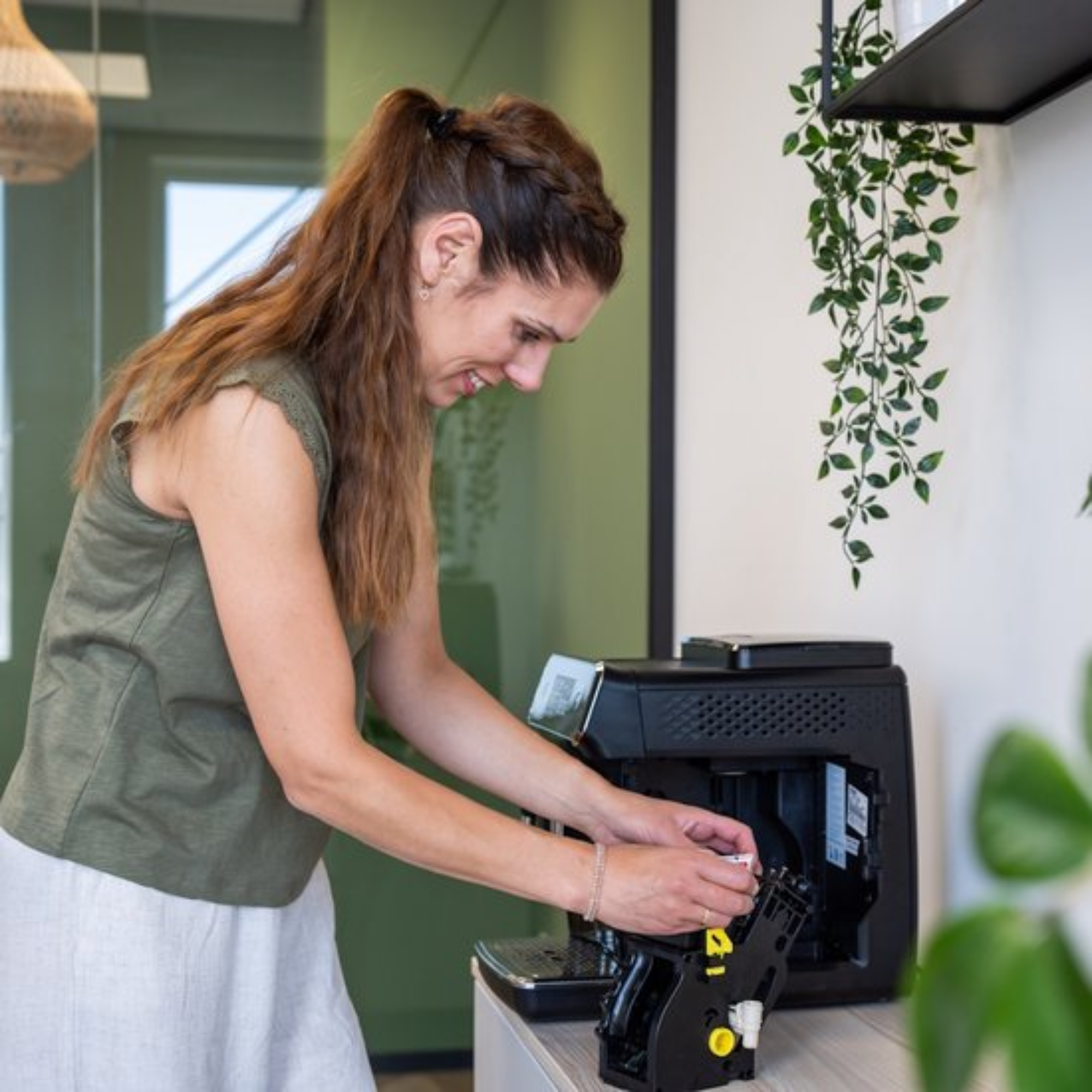 Woman in green top and white skirt fixing a black coffee machine. Plants in the background.