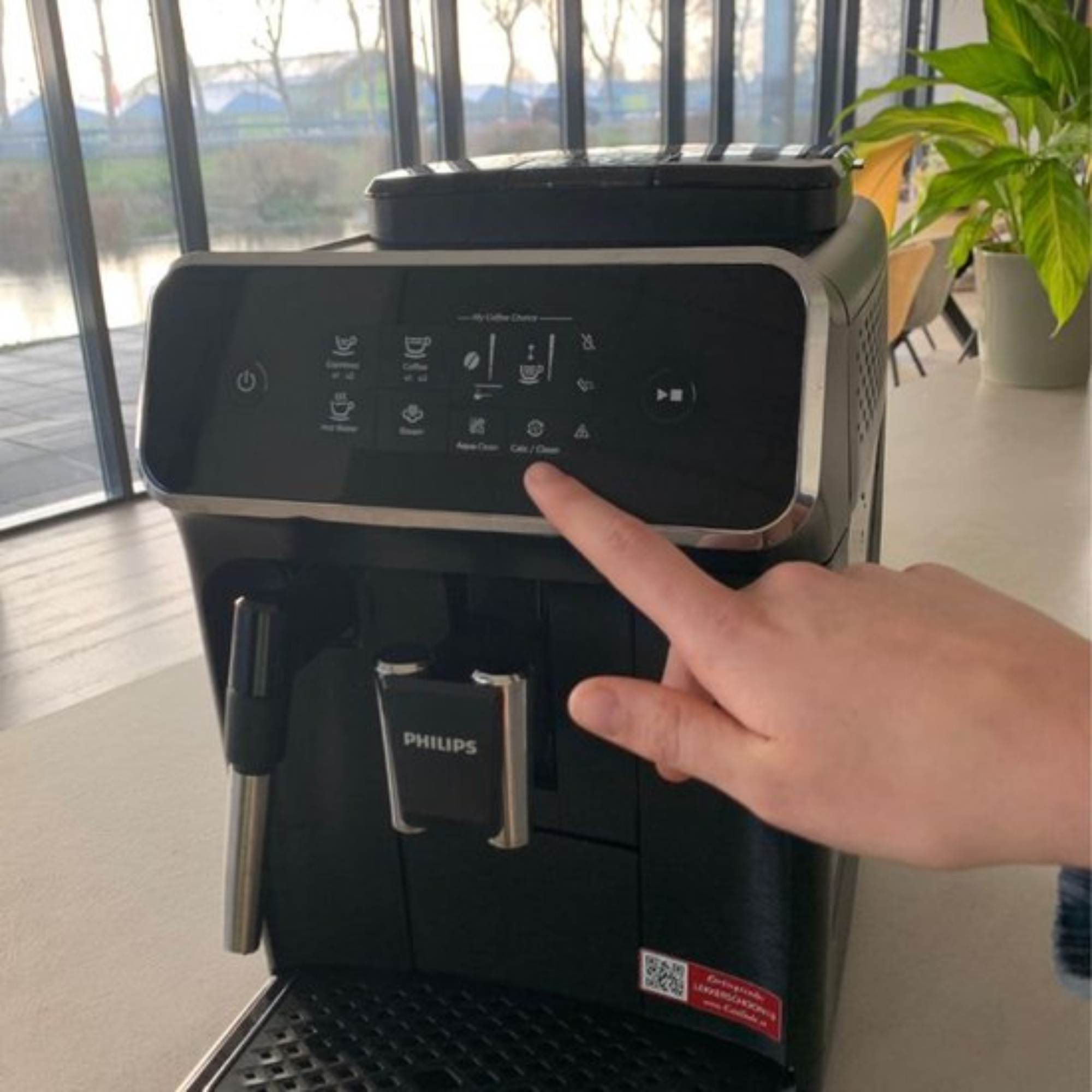A hand presses the Calc/Clean button on a black Philips espresso machine. Background shows a window, trees, and a plant.
