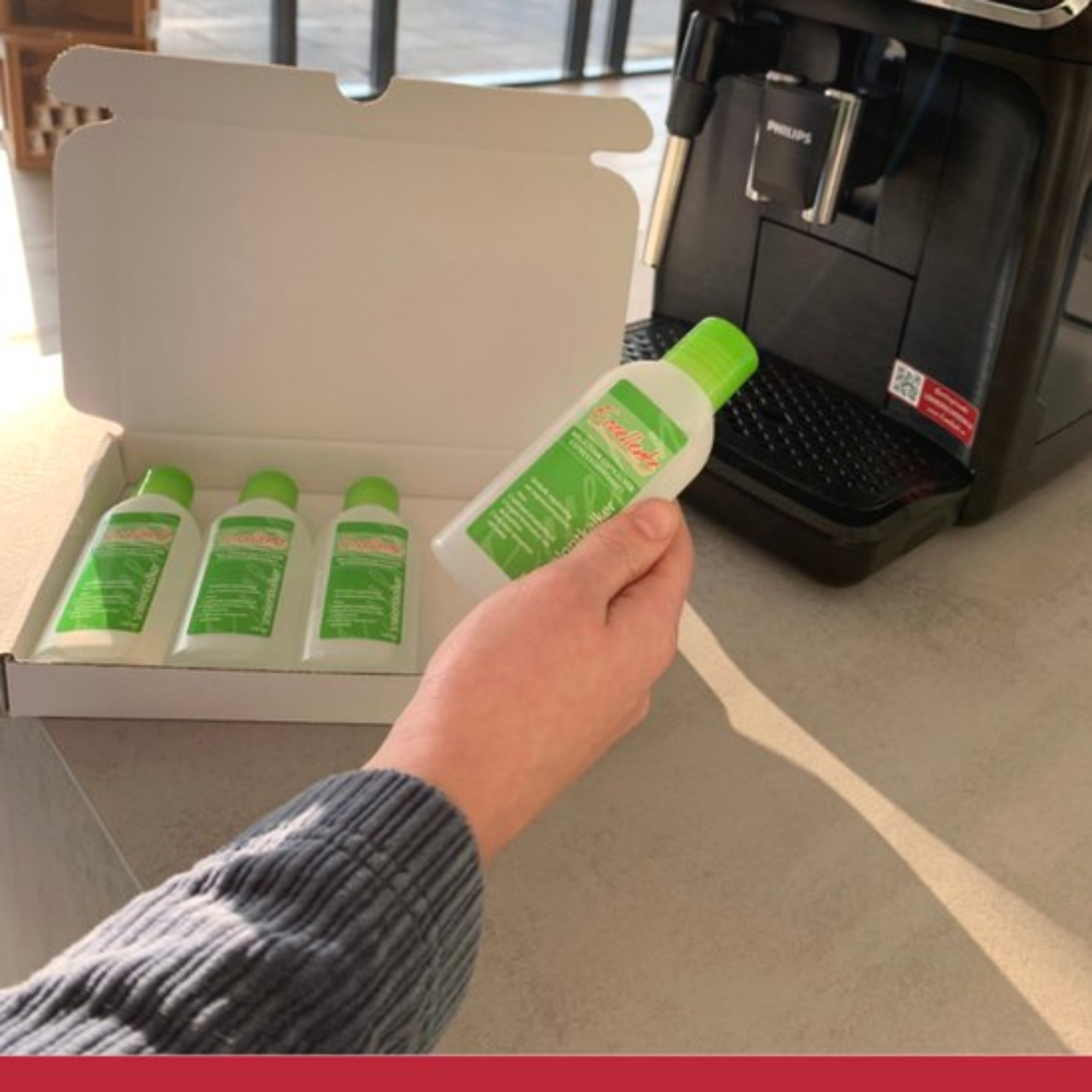 A hand holds a bottle of cleaner. Three more bottles sit in a box. A coffee machine is in the background.