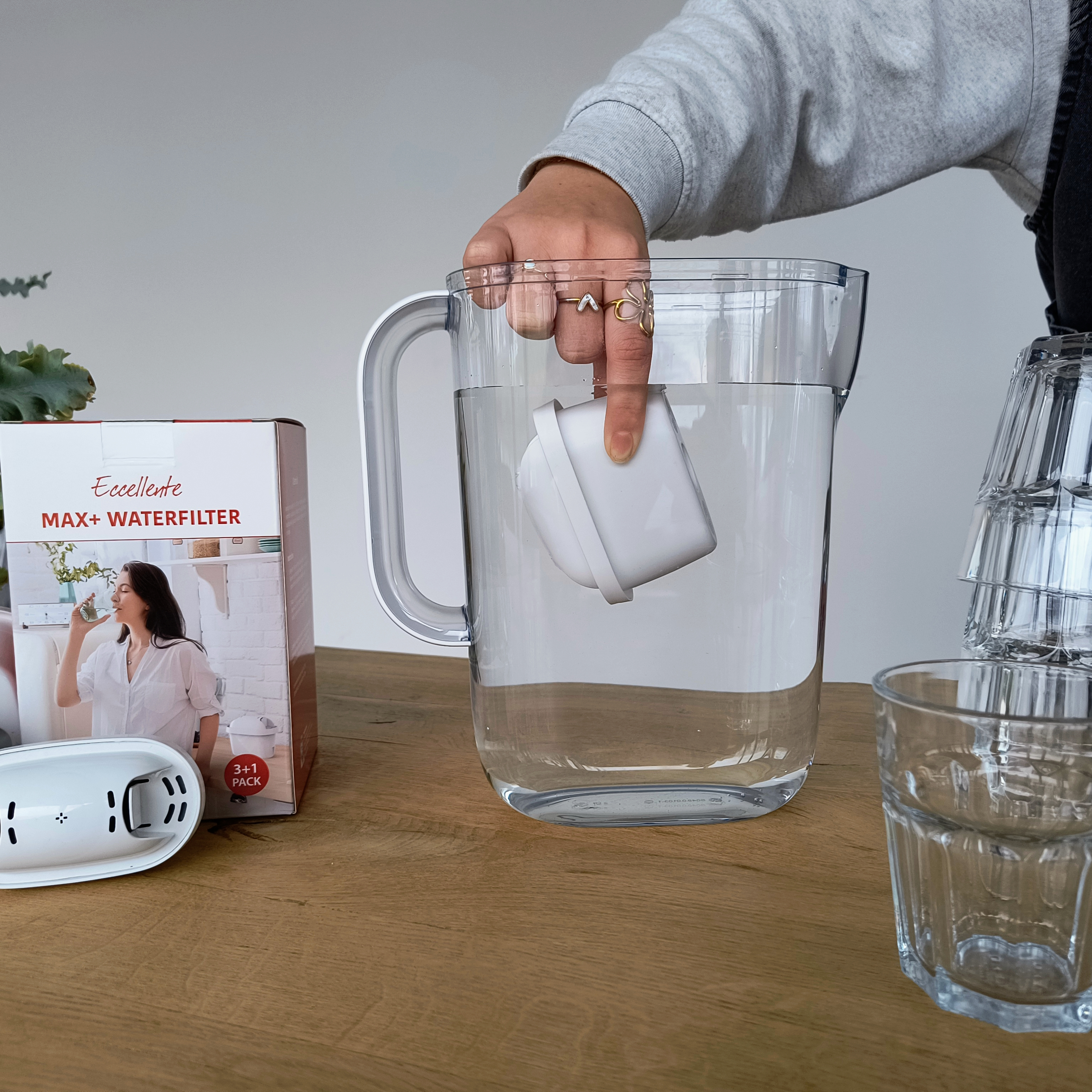A person placing a white filter in a water pitcher. Glasses and a water filter box are on the wooden surface.