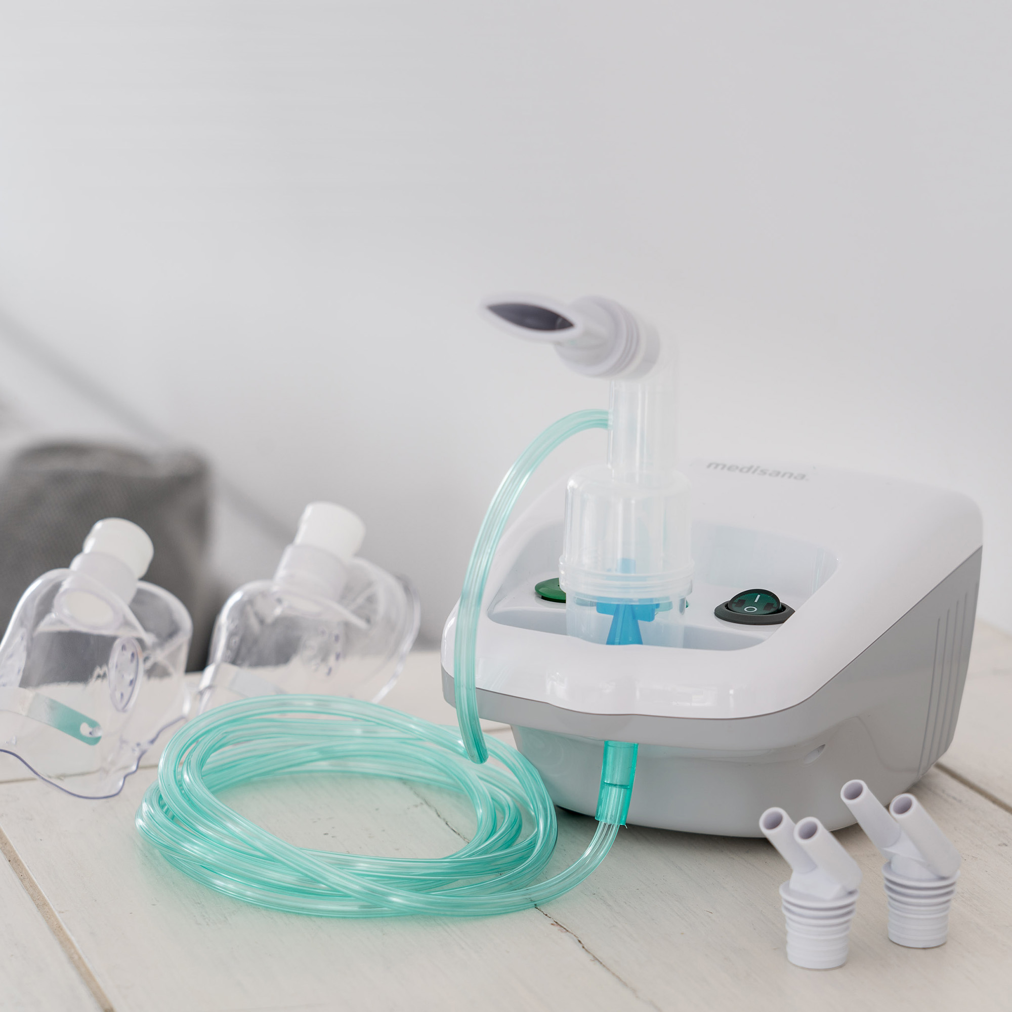 Un nébuliseur blanc et gris avec des tubes et des masques sur une surface en bois blanc. Des accessoires sont également visibles.