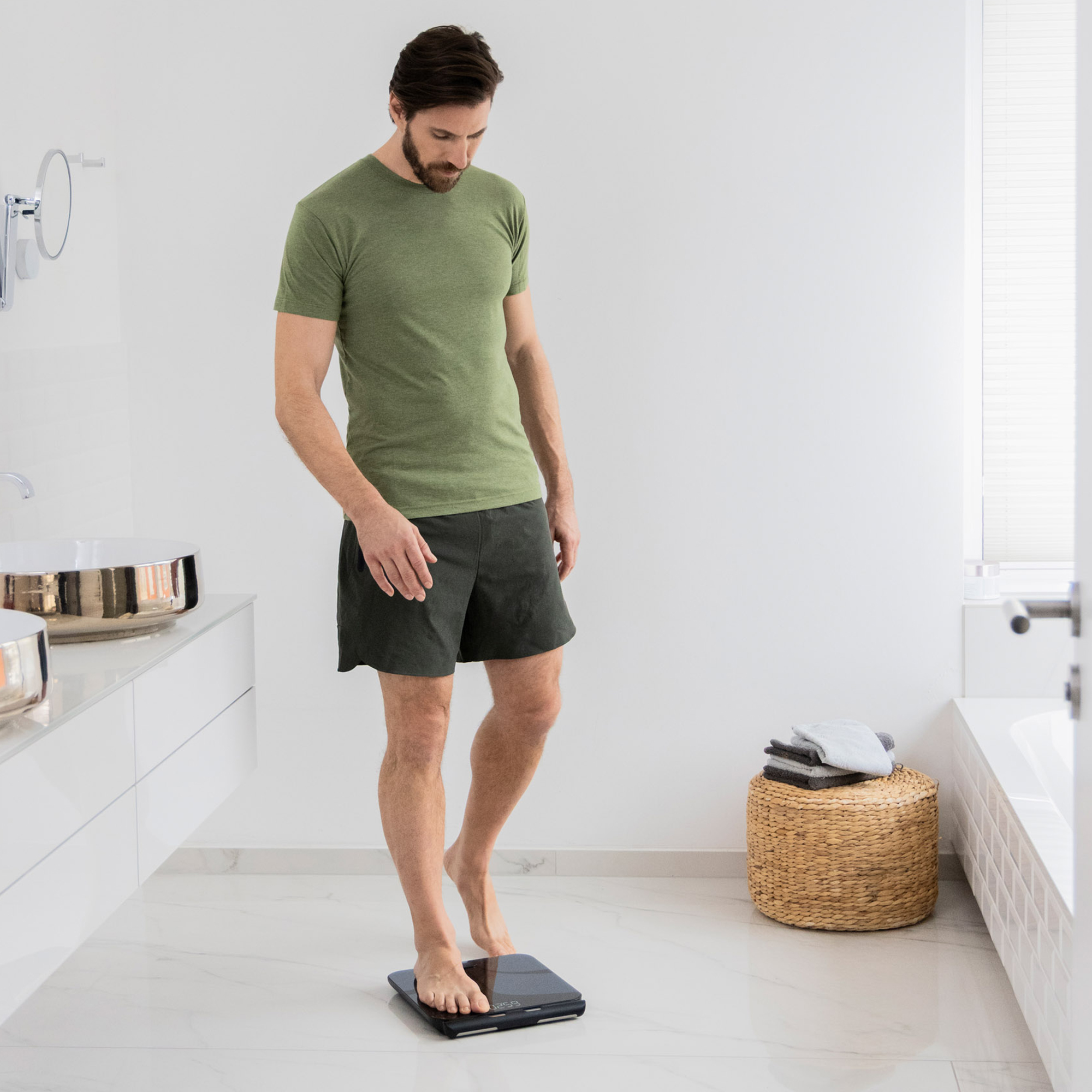 Mann steht auf einer Waage im Badezimmer. Er trägt Shorts und ein grünes Hemd. Hintergrund hat Waschbecken, Handtücher und einen Korntisch.