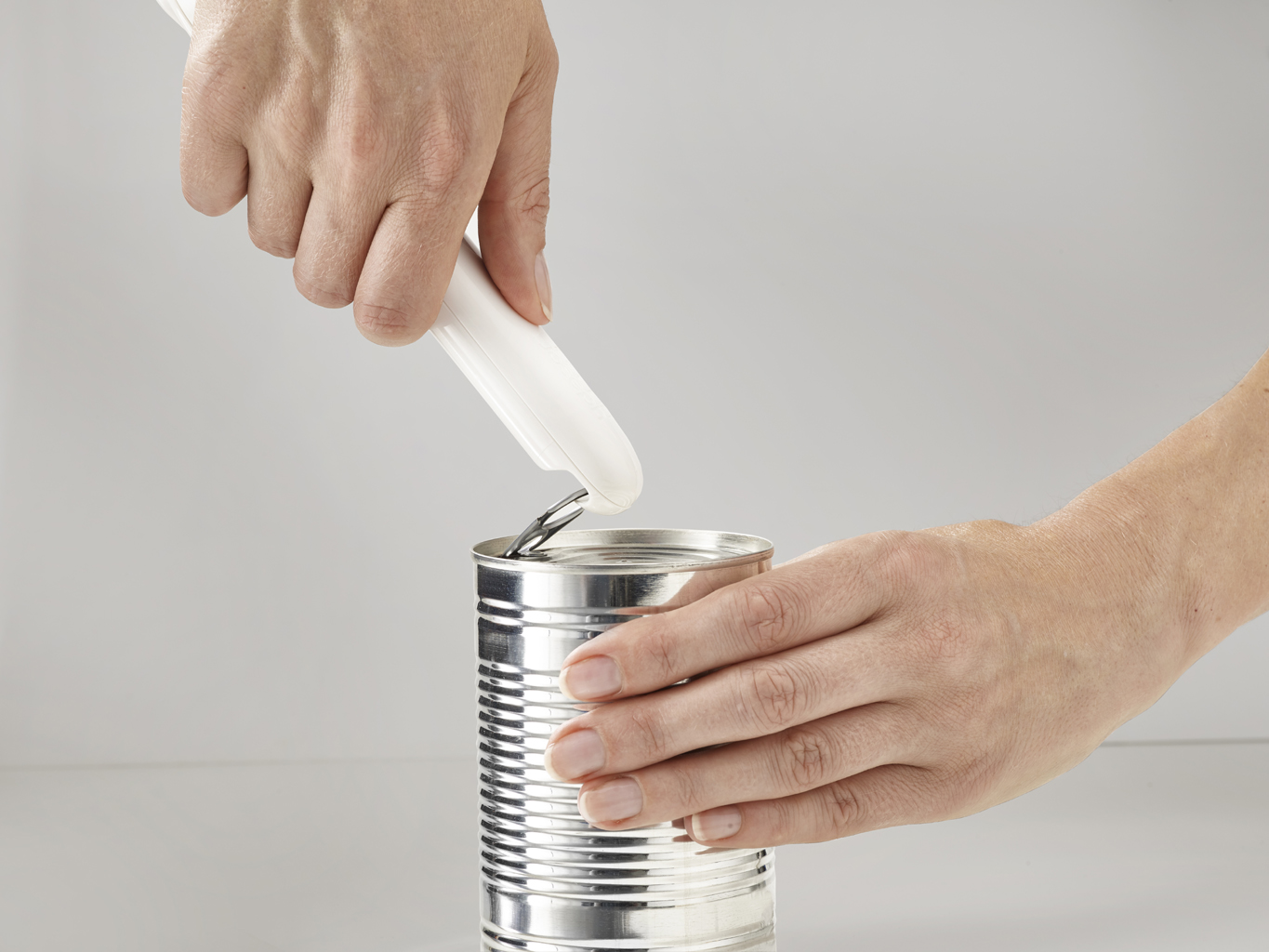 Manos usando un abrelatas blanco para abrir una lata plateada. La lata está sobre una superficie blanca, contra un fondo blanco.