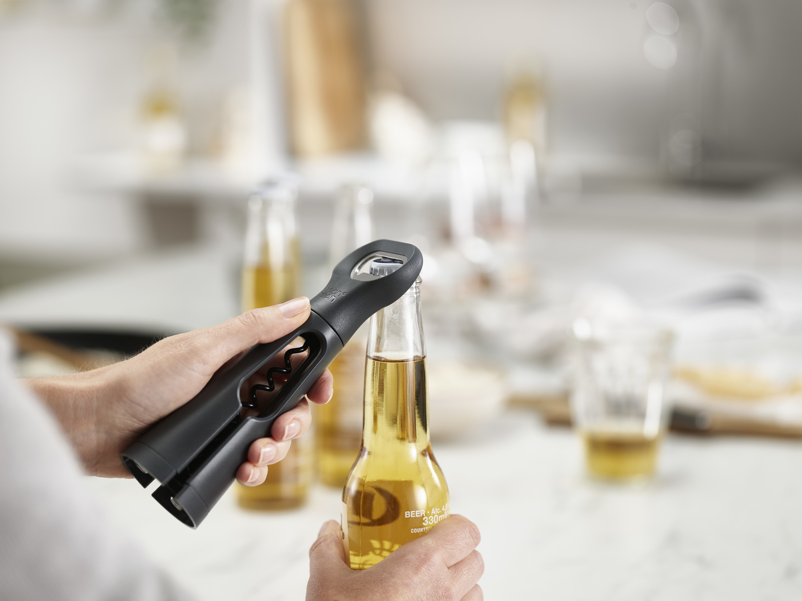 Una persona abre una botella de cerveza con un abridor negro. Otras botellas de cerveza y un vaso están en el fondo borroso.