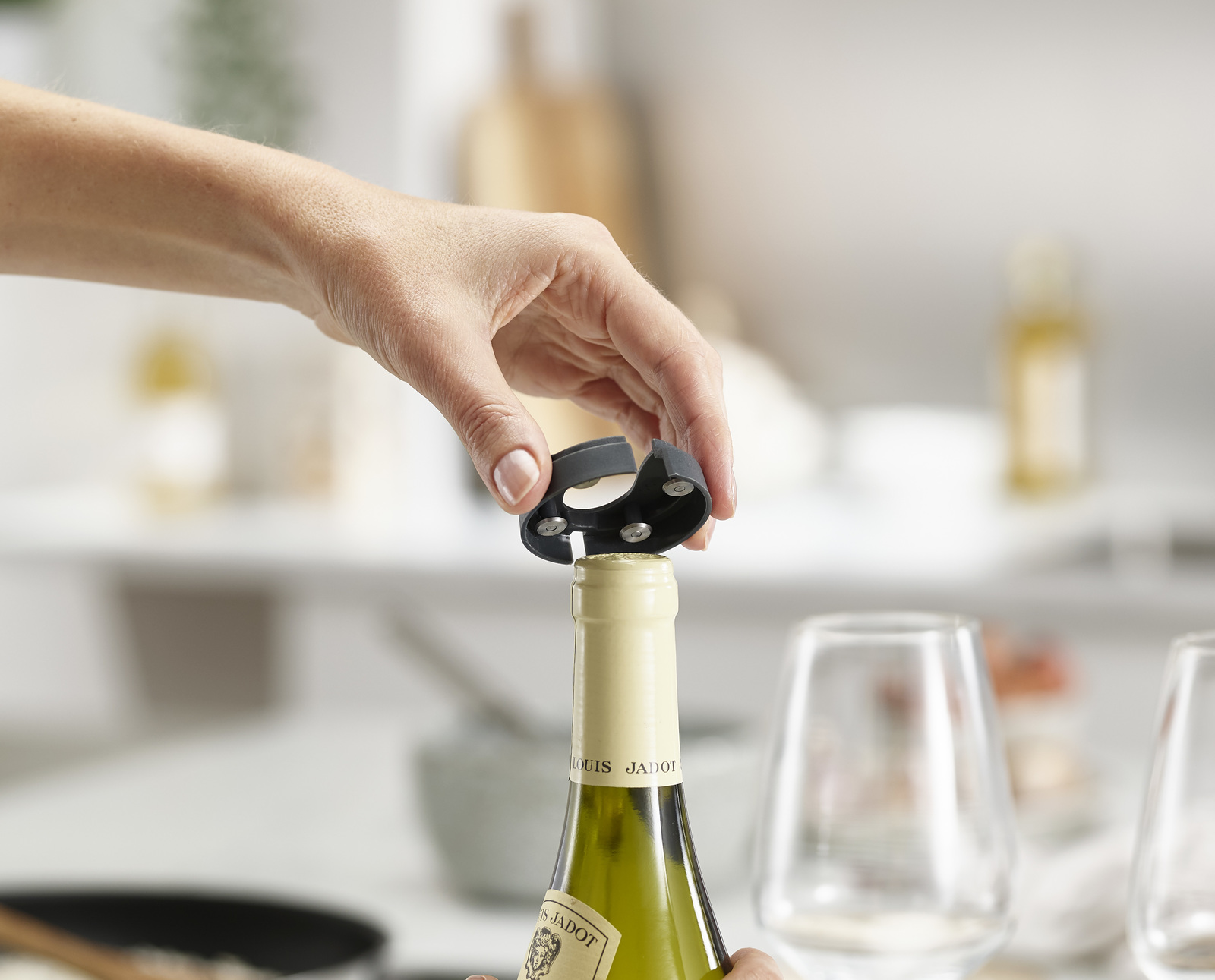 Una mano sostiene un abrebotellas gris sobre una botella de vino. La botella está etiquetada como 'LOUIS JADOT'. Dos copas de vino en el fondo.