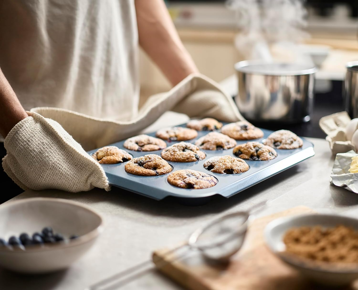 Eine Person hält ein Backblech mit Muffins in Ofenhandschuhen. Dampf steigt aus einem Topf im Hintergrund auf.