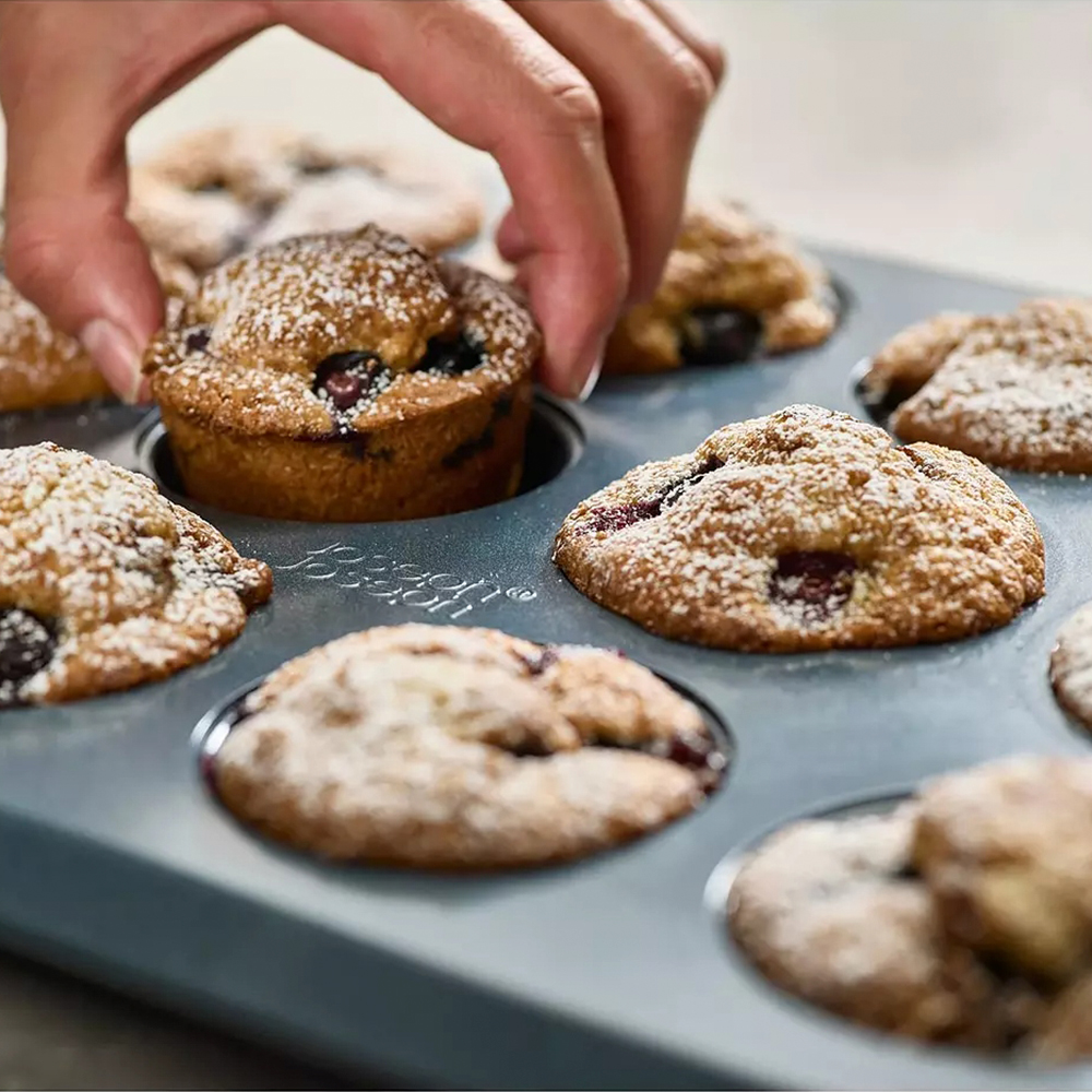 Eine Hand legt einen Blaubeermuffin in eine Muffinform. Weitere Muffins, bestäubt mit Puderzucker, liegen in der Form.