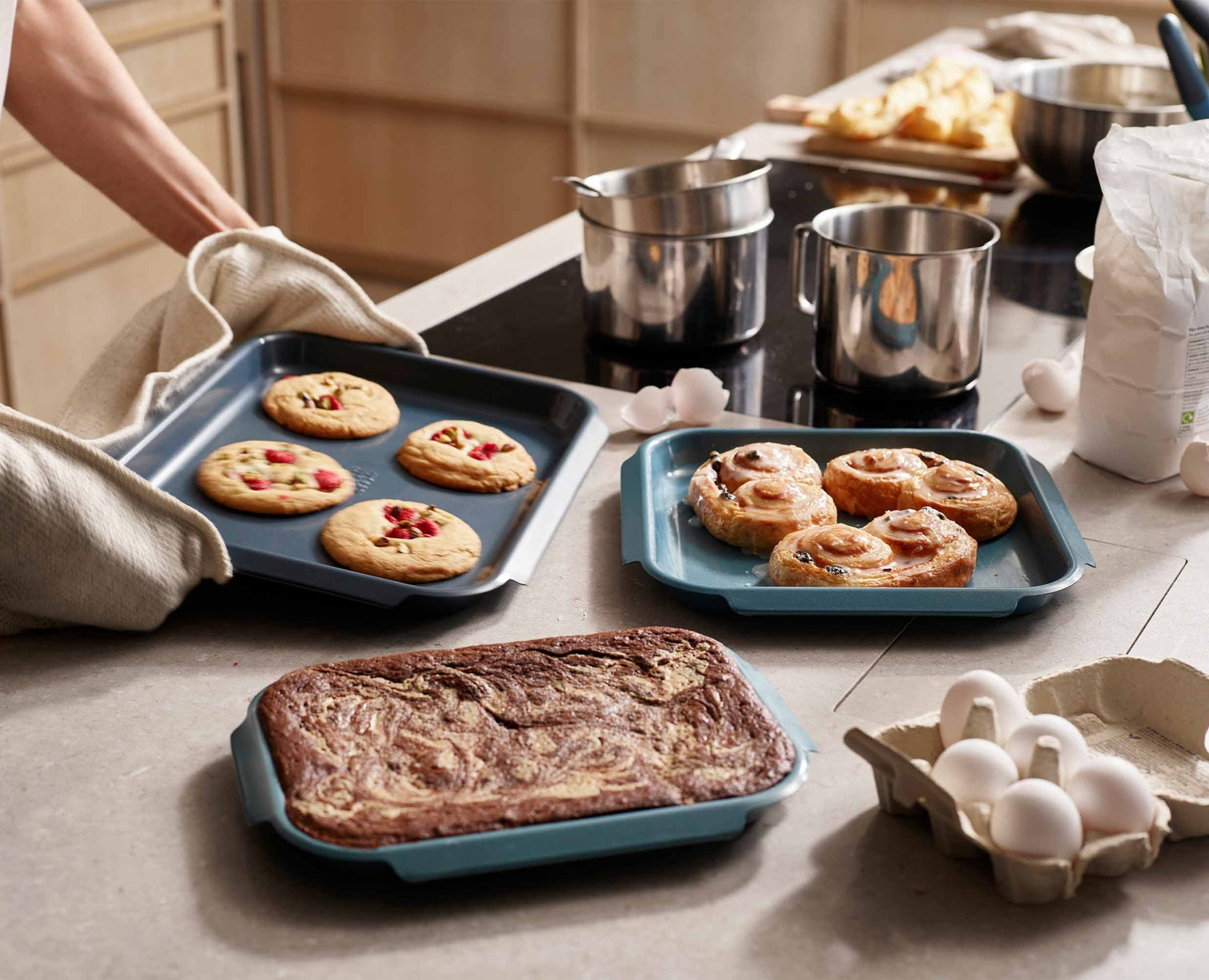 Backszene mit Blechen mit Keksen, Zimtschnecken und einem Brownie. Eier, Mehl und Backutensilien sind ebenfalls vorhanden.