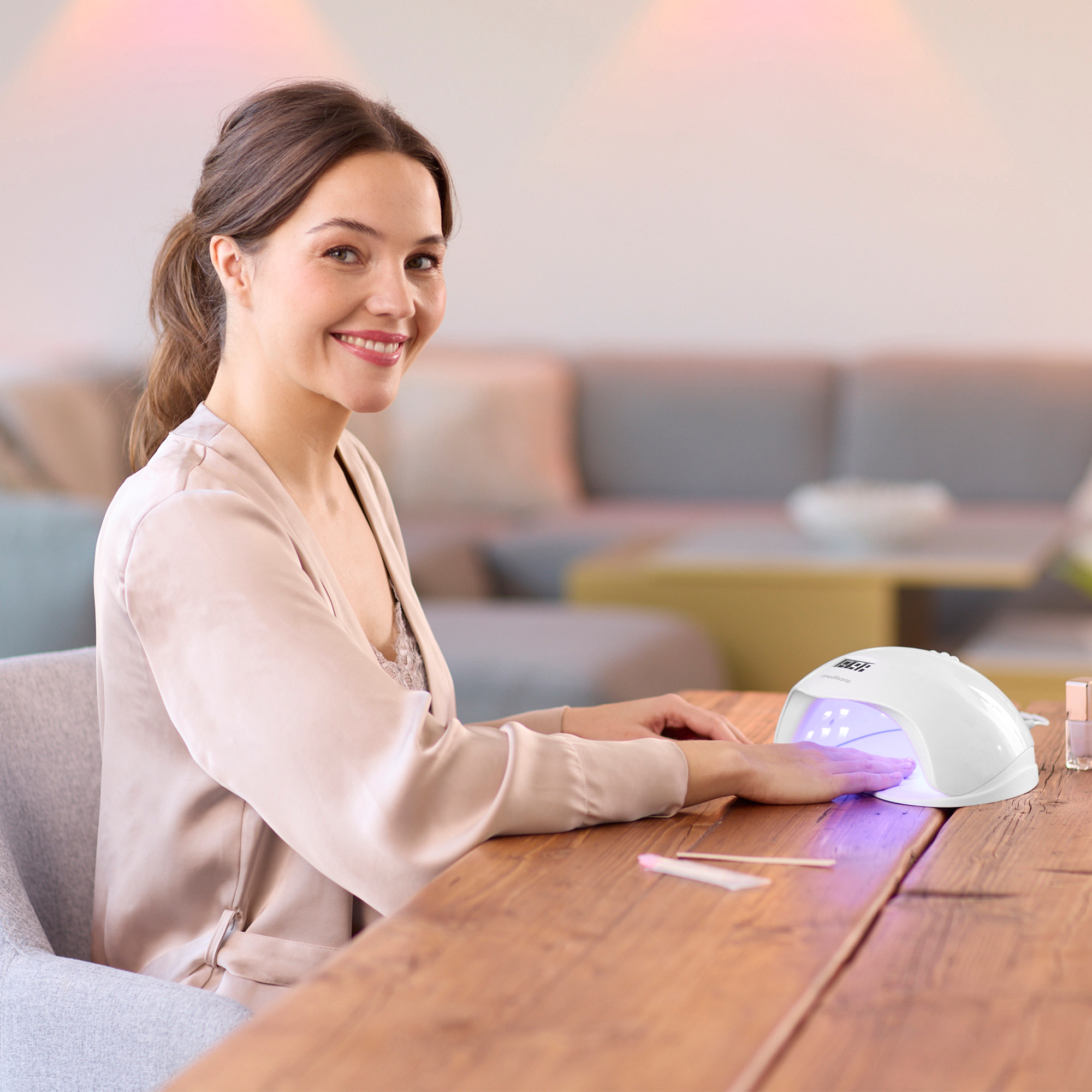 Eine Frau lächelt, während sie sich unter einer UV-Lampe die Nägel macht. Sie trägt einen Bademantel an einem Holztisch. Im Hintergrund ist ein Sofa.