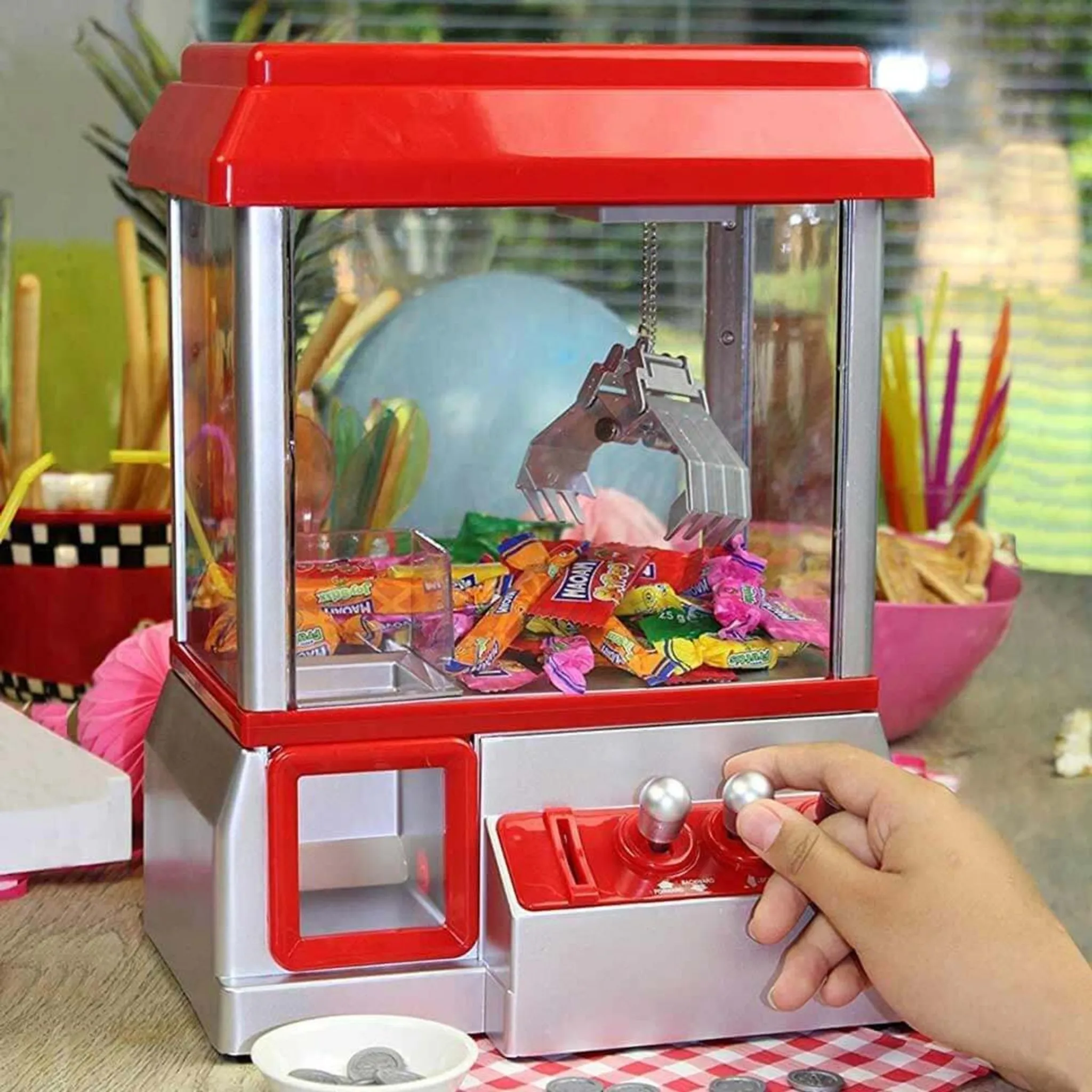 Gezeigt wird ein Greifautomat. Eine Hand bedient den Joystick. Im Inneren befinden sich bunte Süßigkeiten, ein Greifer und ein roter Deckel.
