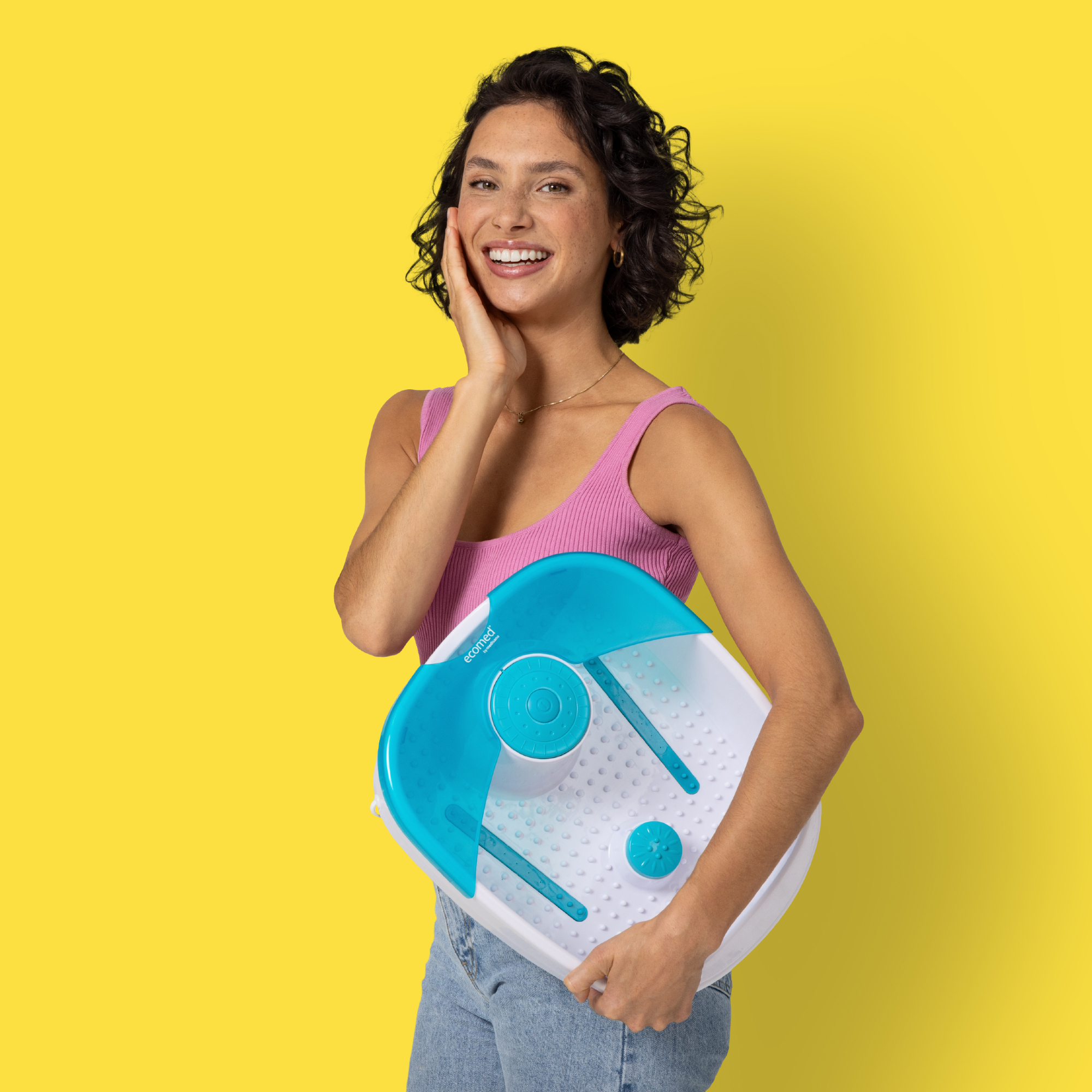 Una donna tiene un pediluvio, sorridendo. Indossa un top rosa e jeans, su sfondo giallo.