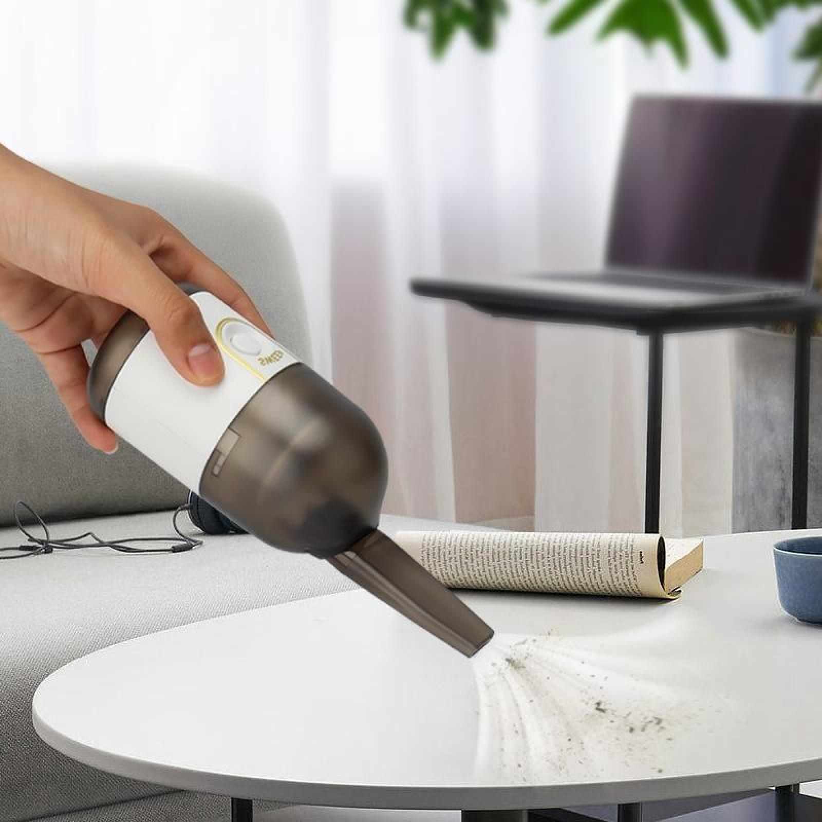 Eine Hand hält einen weiß-grauen Handstaubsauger über einem weißen Tisch und reinigt Staub. Ein Laptop und ein Buch sind sichtbar.