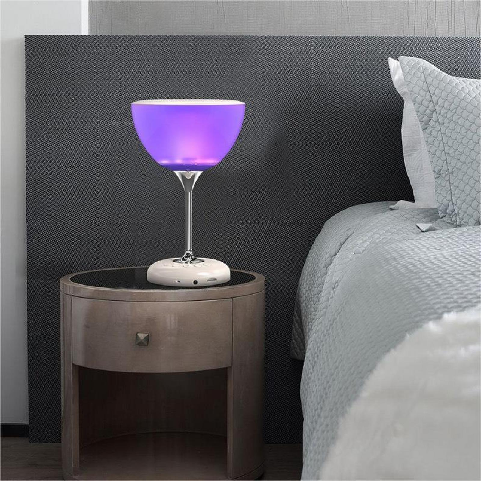 Ein Nachttisch mit einer lampe in Form eines Weinglases. Die Lampe ist lila. Im Hintergrund ist ein Bett.