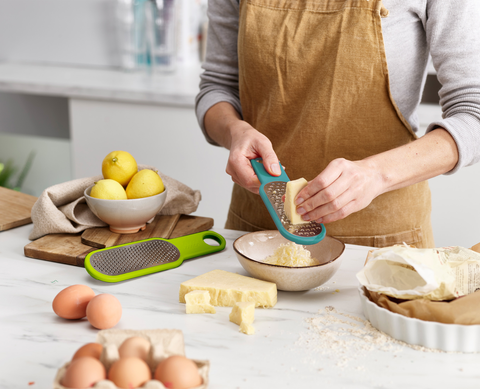 Una persona grattugia il formaggio. Limoni, uova e formaggio sono sul bancone. Sono visibili una grattugia e una ciotola con formaggio grattugiato.